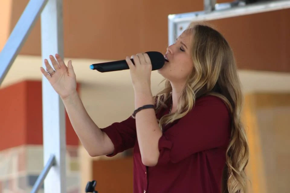 Blonde teenager in a red shirt singing passionately into a microphone. The background is different shades of orange