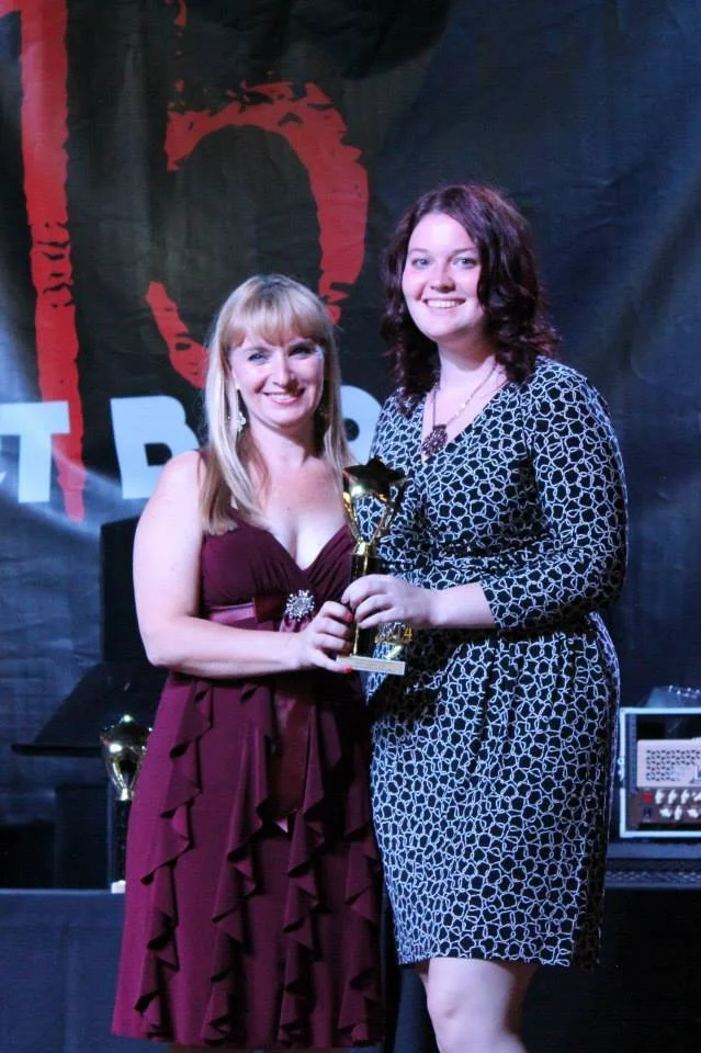 Blonde woman in a red dress and brown haired woman in a black and white dress holding a trophy together at the 2014 WCCMA award show