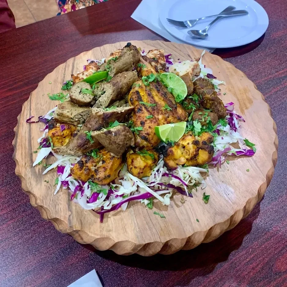 A large wooden serving platter with grilled chicken, skewered beef, and other grilled meats on a bed of shredded cabbage and garnished with lime wedges and cilantro. The platter is on a dark wooden table, with a plate, spoon, and fork in the backgrou