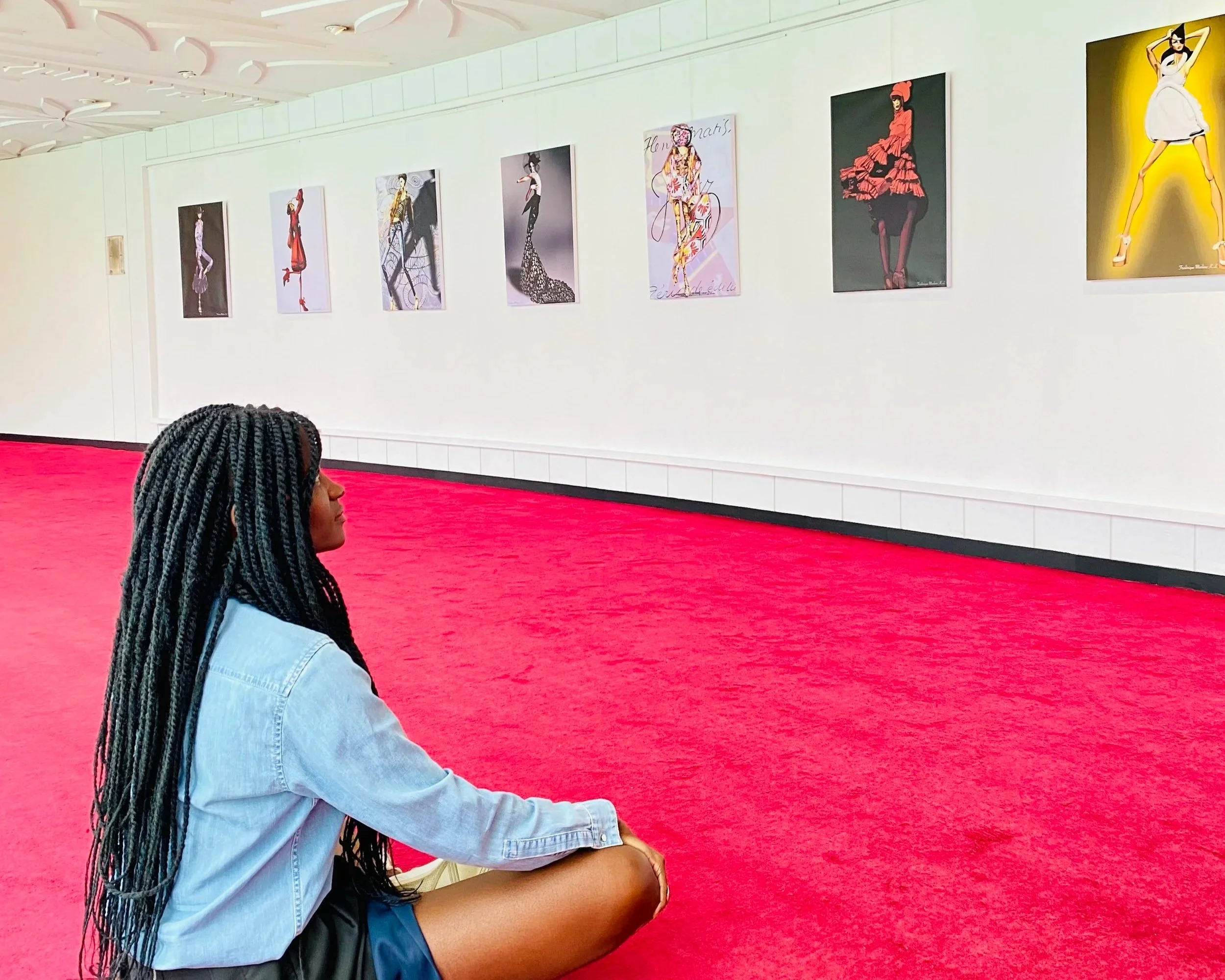 Frédérique NANAN, assise par terre dans une galerie d'art, regardant des tableaux de mode suspendus au mur blanc. Le sol est rouge vif. Sofitel Abidjan