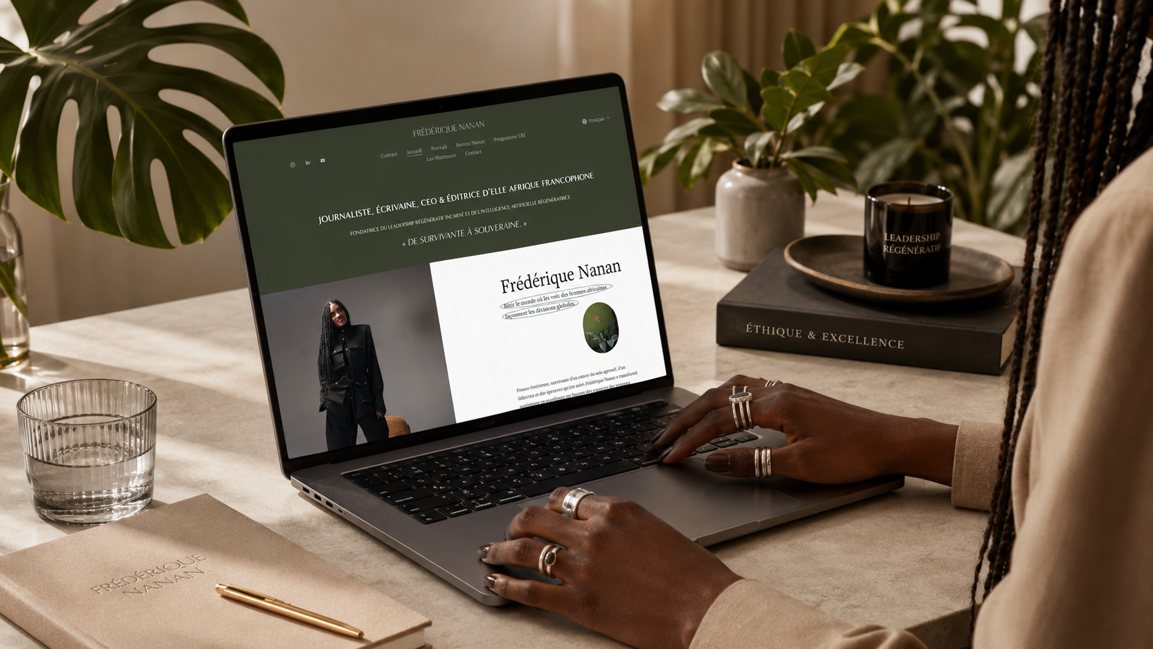 Une personne utilisant un ordinateur portable sur une table, affichant un site web personnel de Frédérique Nanan, avec des plantes décoratives, un livre intitulé 'Éthique & Excellence', une bougie, un carnet beige avec un stylo doré, et un verre d'eau.