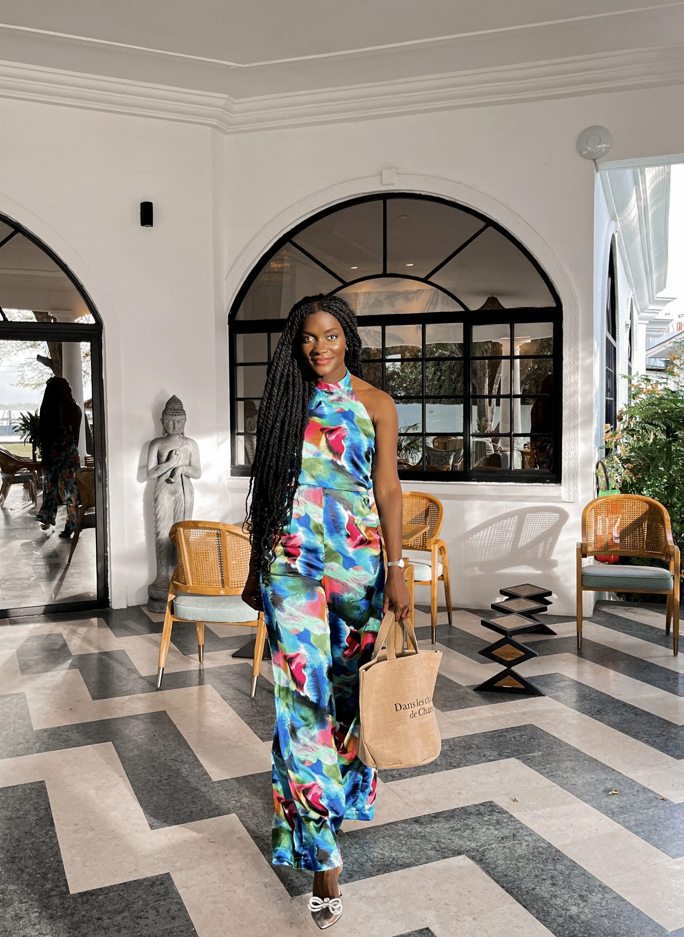 Femme debout dans une salle lumineuse portant une robe colorée et tenant un sac en toile beige.