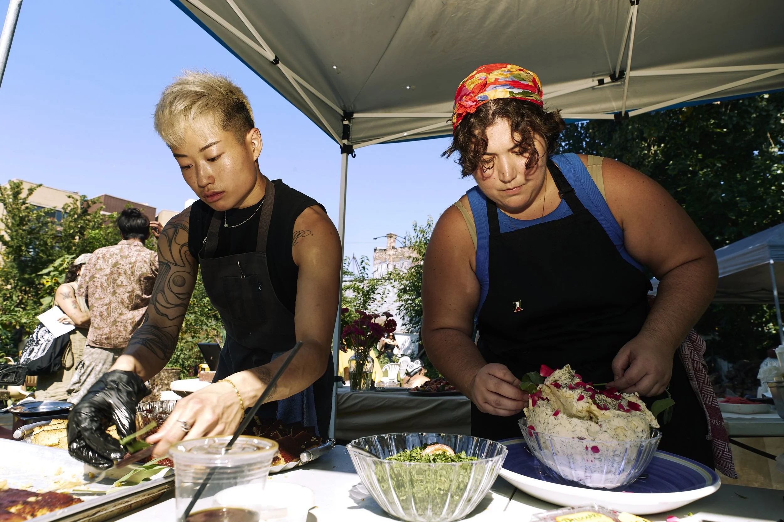 Two people preparing food at an outdoor event under a tent. One is slicing a dish, and the other is mixing a large bowl of salad or dessert with rose petals on top.