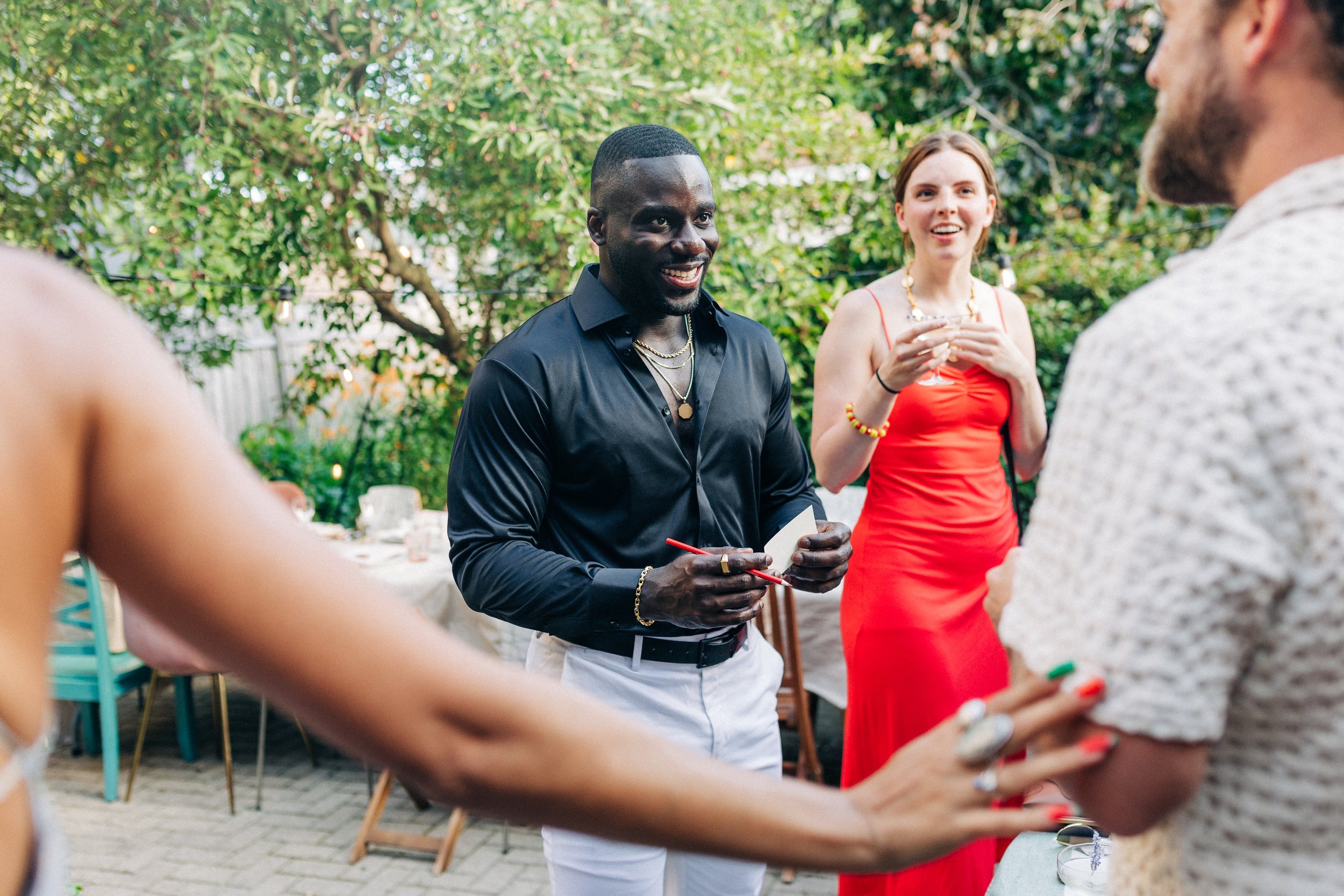 People socializing outdoors, including a man in a dark shirt and white pants, and a person in a red dress, with others partly visible, set in a lush garden area with trees and string lights.
