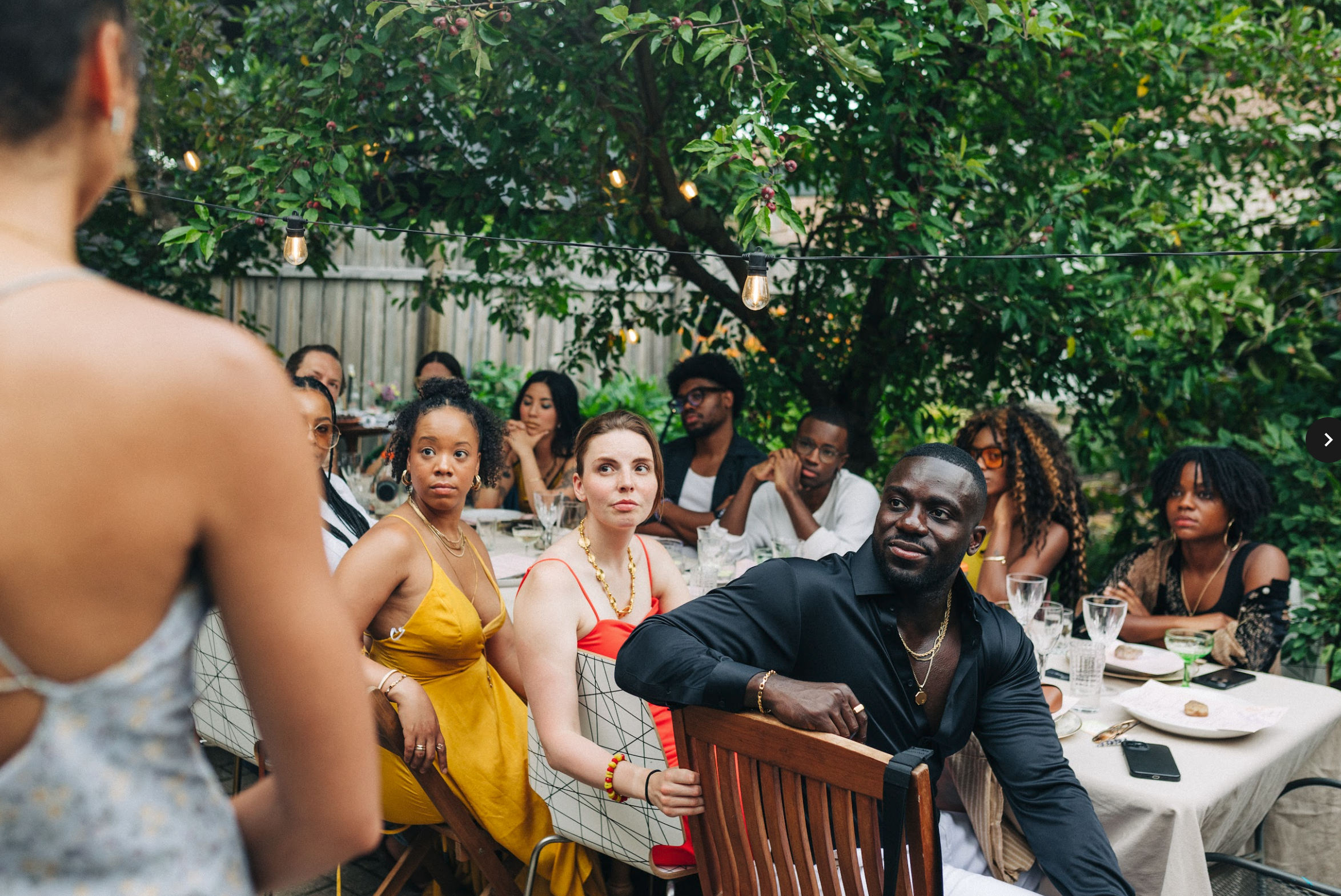 A group of people sitting at a long table turn around to look at a person speaking in the foreground