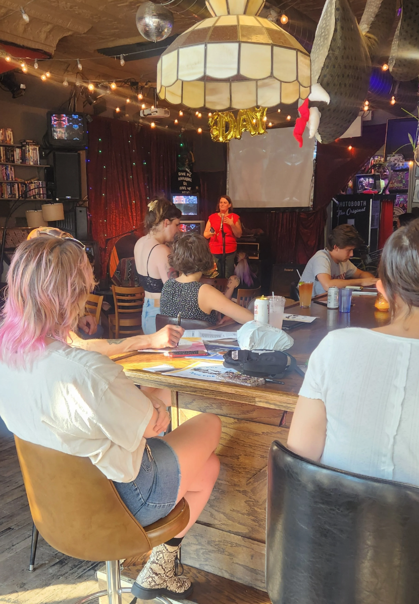a person on stage is speaking in front of five people in a bar setting. The photo is from far away and we're looking at the backs of people.