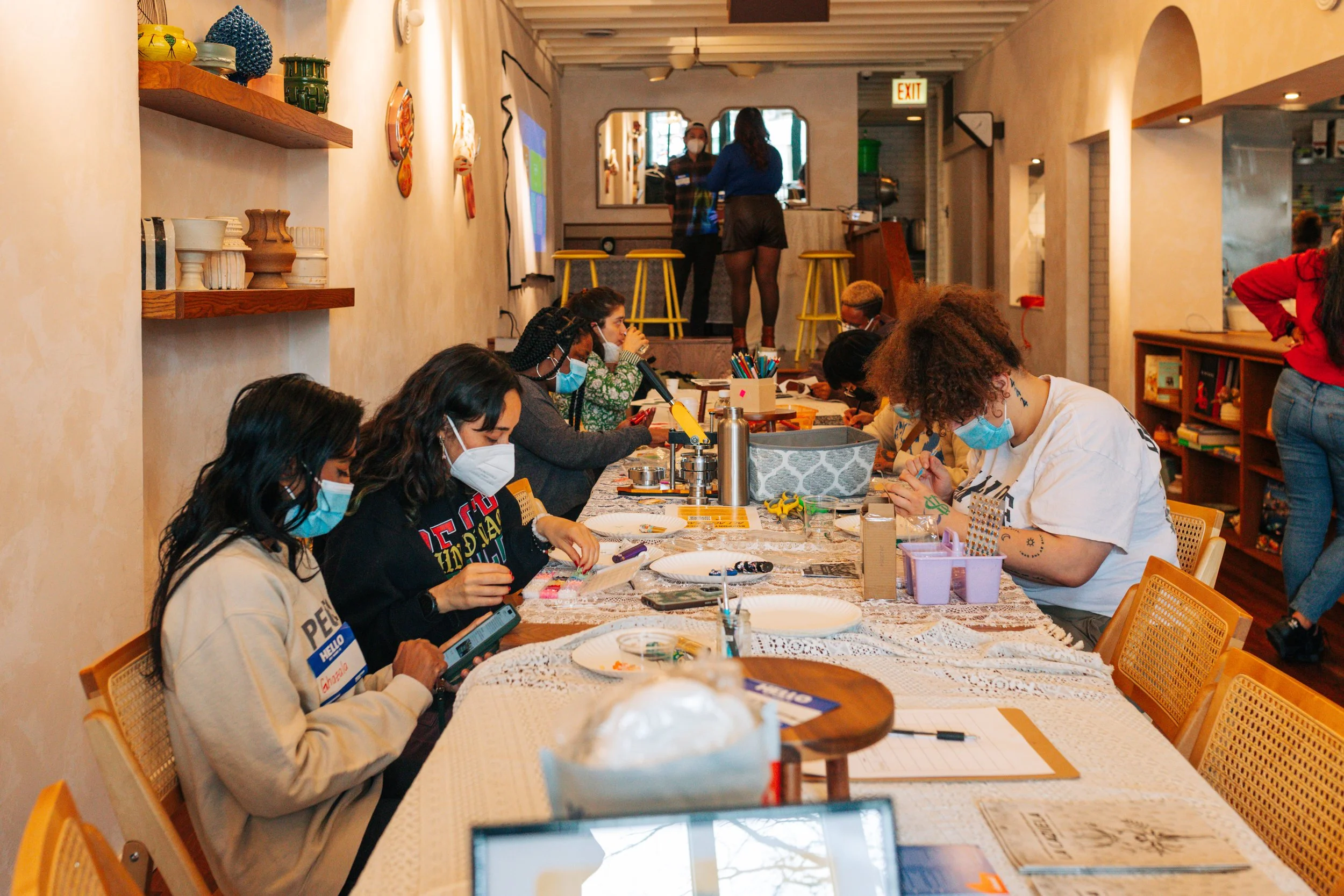 People sitting at a long table engaging in arts and crafts activities inside a cozy cafe or workshop space, with shelves and people standing in the background.