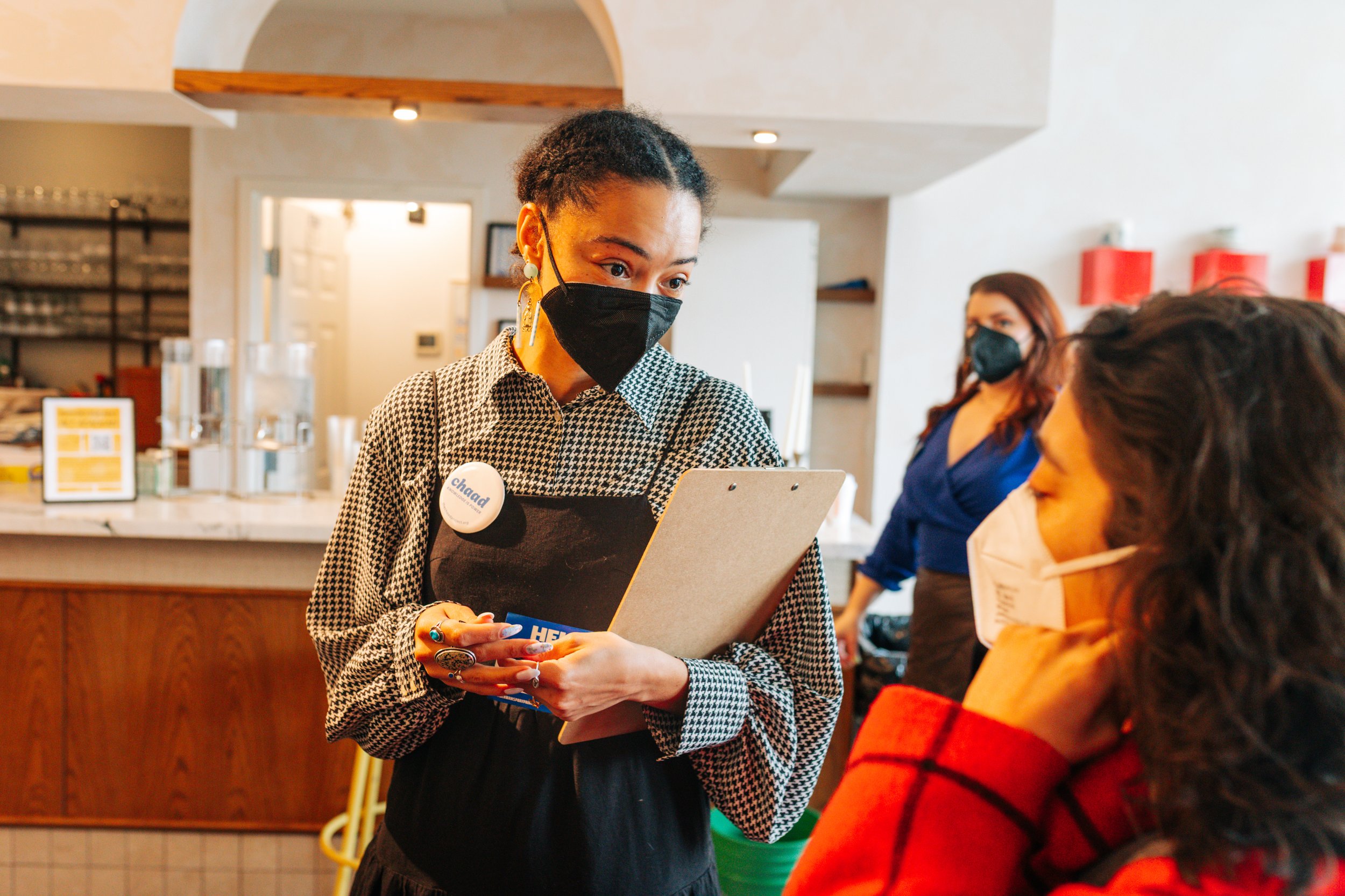 A pers with short hair and a patterned shirt standing at a counter, holding a clipboard and interacting with a customer in a restaurant or café. Both peopleare wearing masks, and there is another perso in the background wearing a blue shirt and mask.