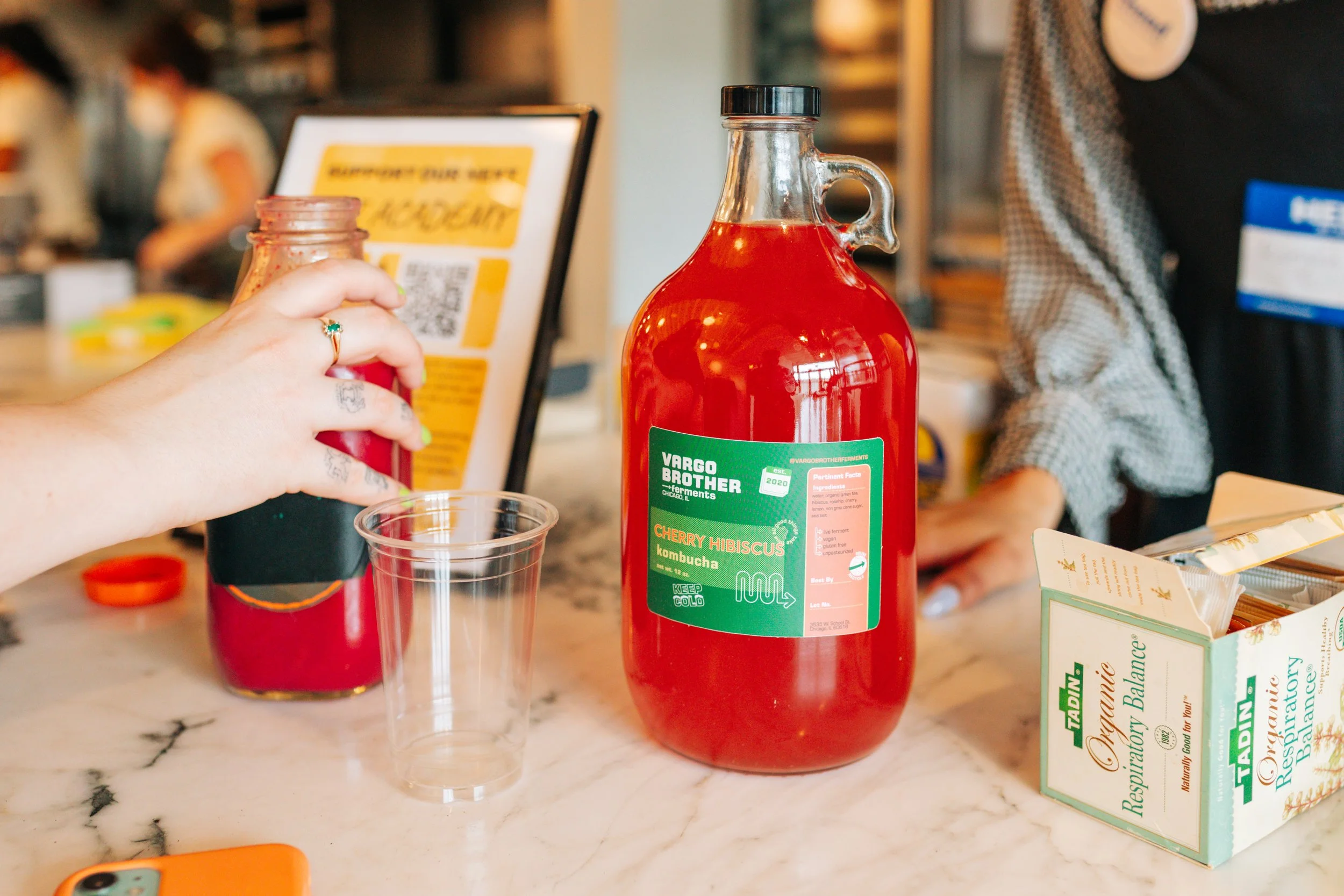 A person pouring kombucha from a large red bottle labeled 'Vargo Brother Ferments Cherry Hibiscus Kombucha' into a clear plastic cup, with a box of organic respiratory balance tea on the countertop.