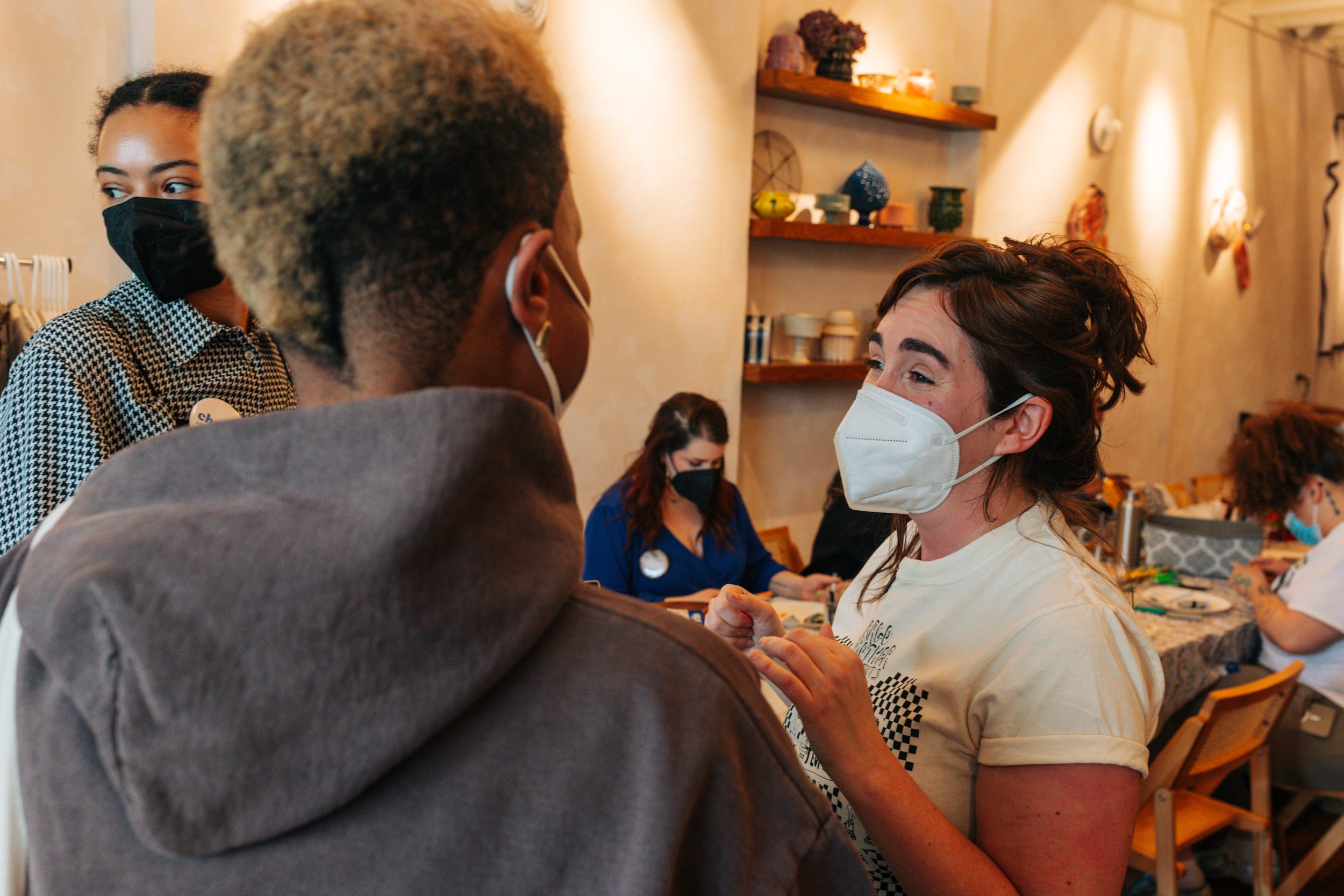 A person smiling and talking to another, both wearing masks, in a cozy indoor space with shelves and other people in the background.