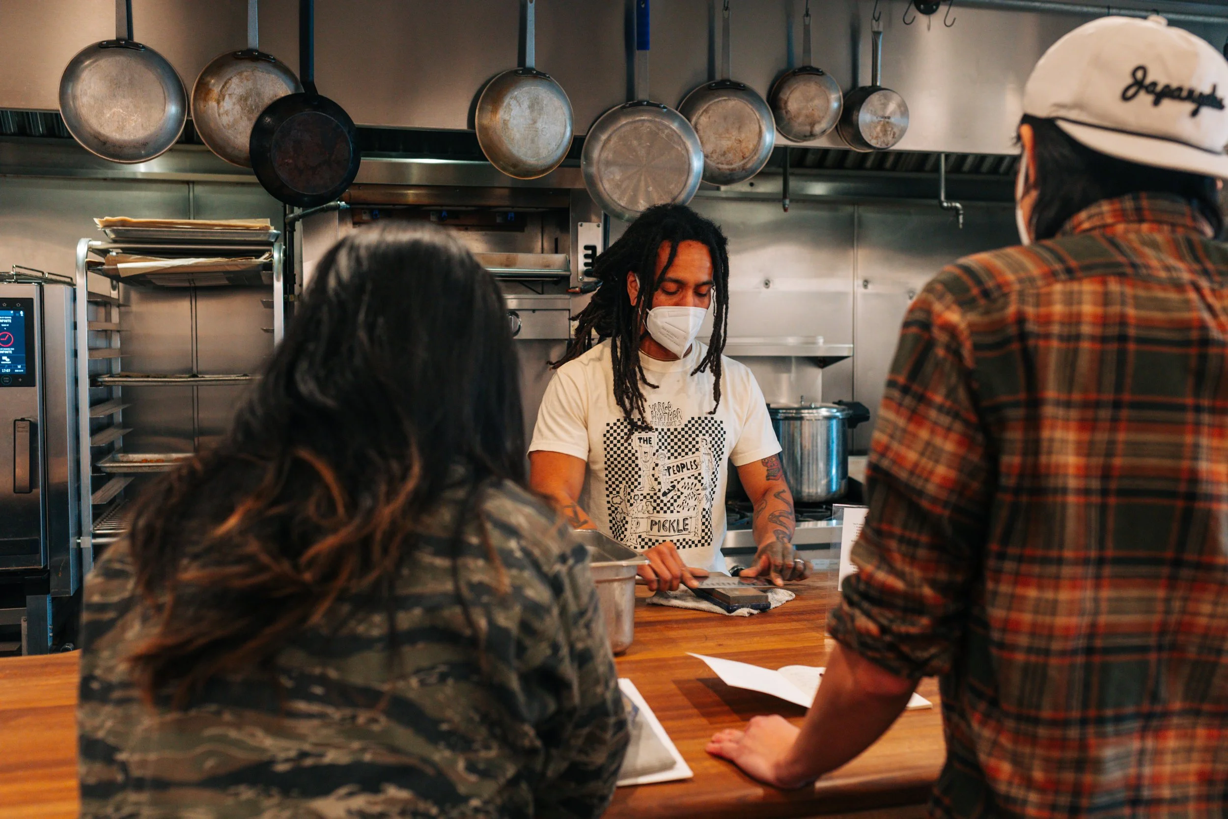 Three people inside a kitchen with hanging pots above, one person with tattoos and dreadlocks wearing a face mask and a graphic T-shirt is cutting something on a cutting board, two others are standing at a counter and looking at her, with papers and a book in front of them.