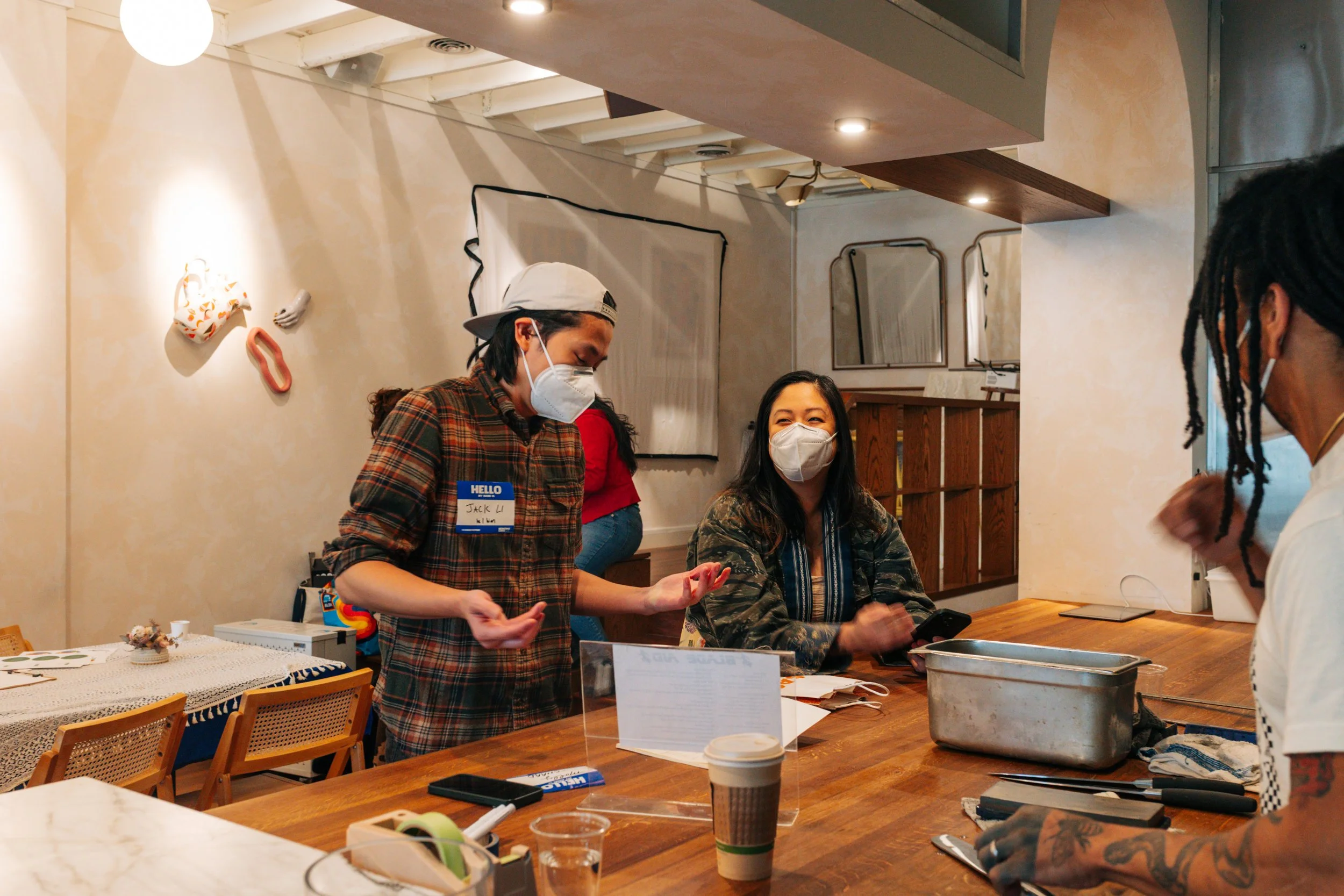 Three people wearing masks standing and sitting at a wooden counter inside a cafe or restaurant, with one person in a plaid shirt and a name tag talking to another person with dreadlocks.