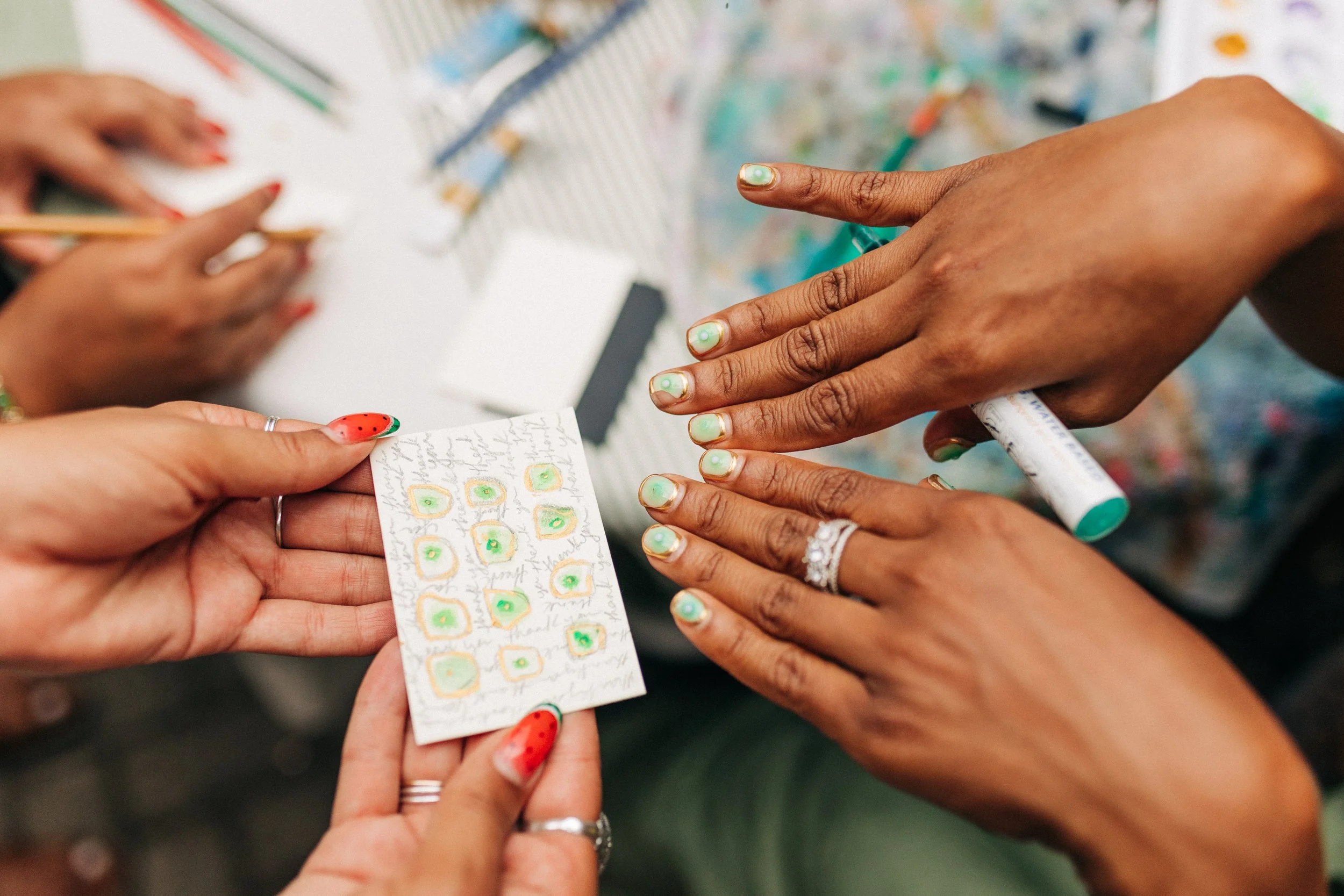 Close-up of hands displaying nail art, with a nail polish pen in one hand and a sheet with nail designs in the other, amid a colorful nail polish palette and manicure supplies.