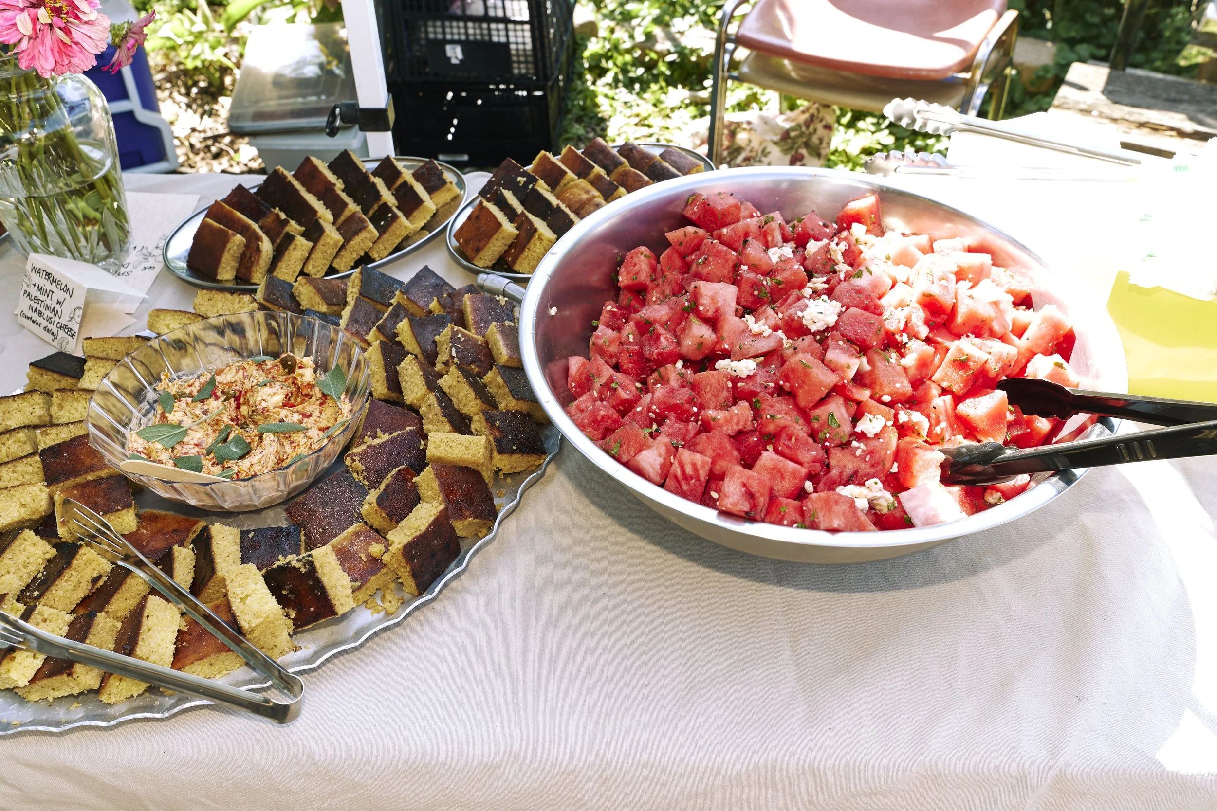 Trays of sliced cake, a bowl of watermelon salad, and a vase of flowers on a table outdoors.