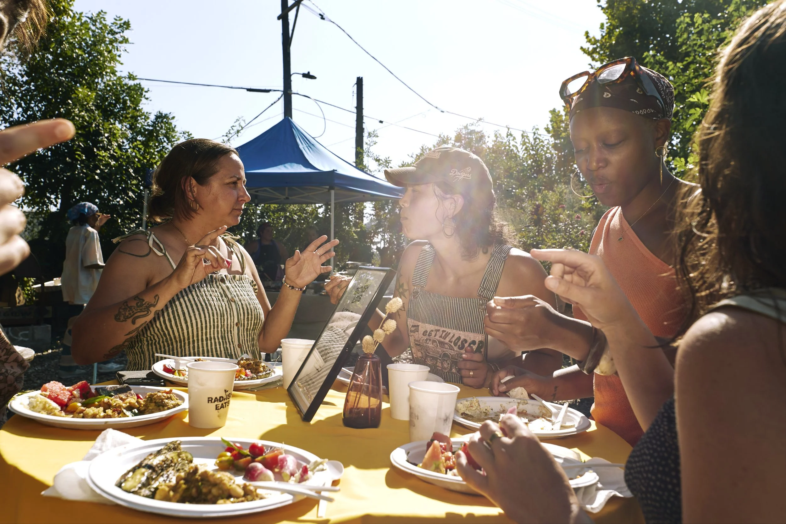 People enjoying a meal at an outdoor gathering around a yellow table, with various dishes and drinks, engaged in conversation, under a bright sunny sky with tents and trees in the background.