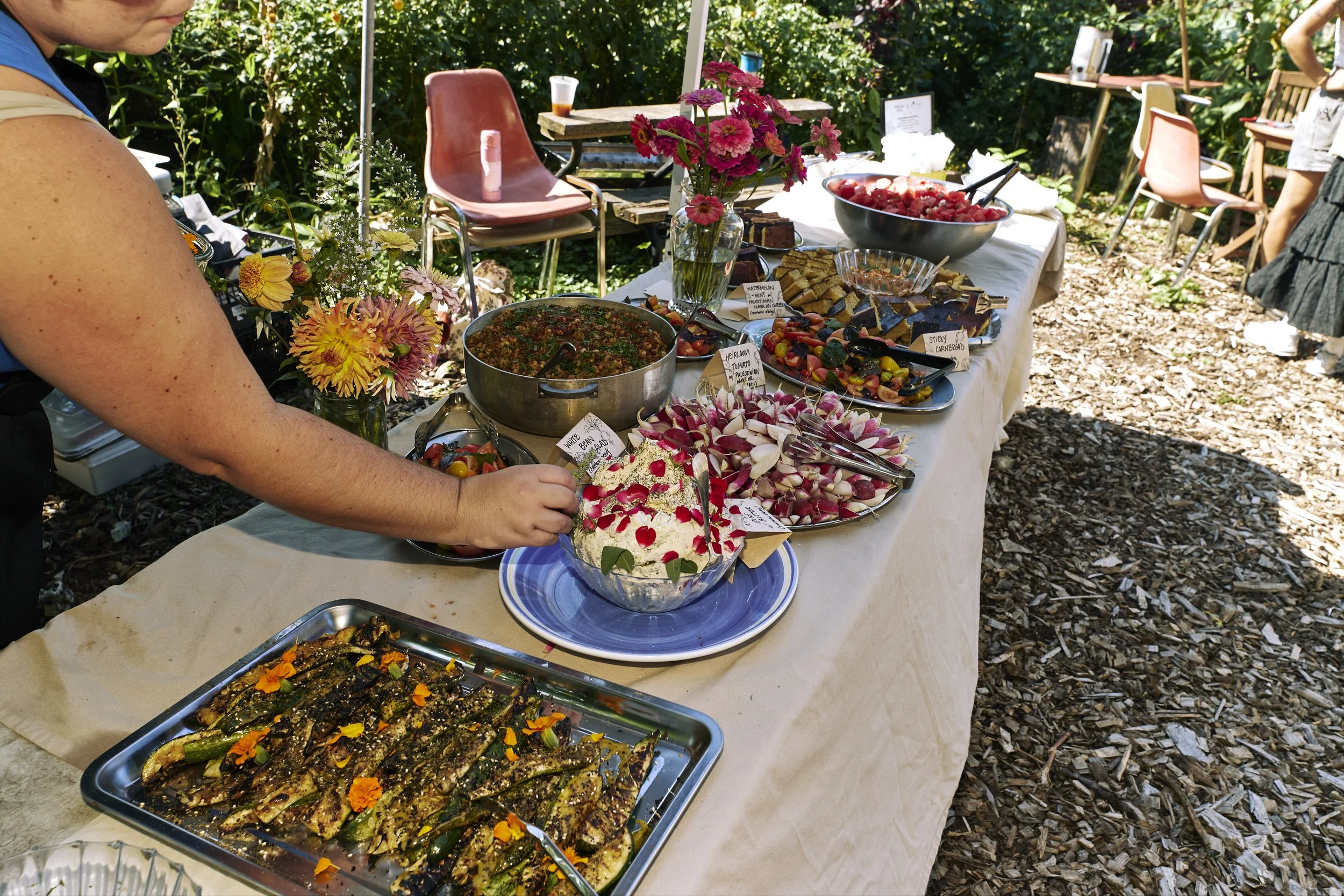 A buffet table outdoors with various dishes, including a large salad with radishes, grilled vegetables, fruit, and desserts, decorated with flowers.