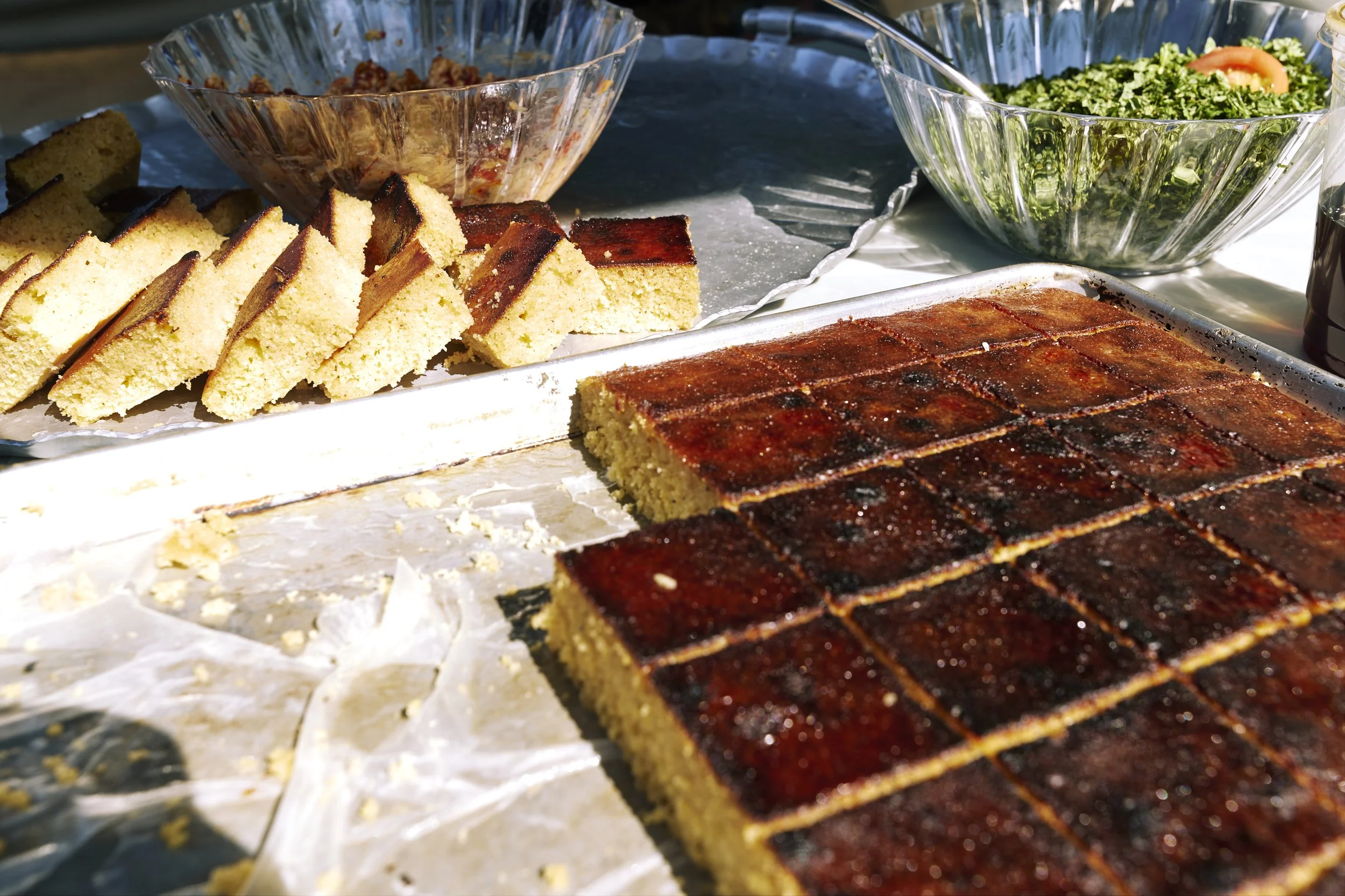 Slice of yellow cake with a dark brown top in a large rectangular pan, surrounded by crumbs, and whole slices of the same cake, along with bowls of salad with greens and vegetables.