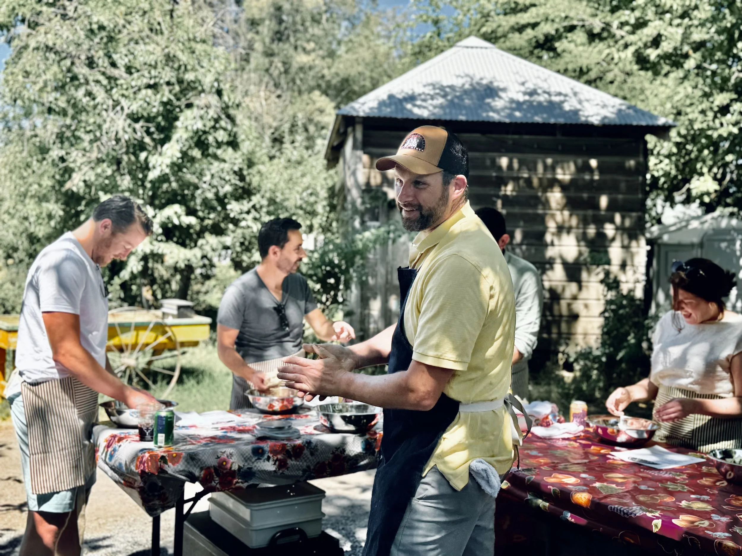 Group of people cooking outdoors at a table with bowls and ingredients, surrounded by trees and a wooden shed, sunny day.