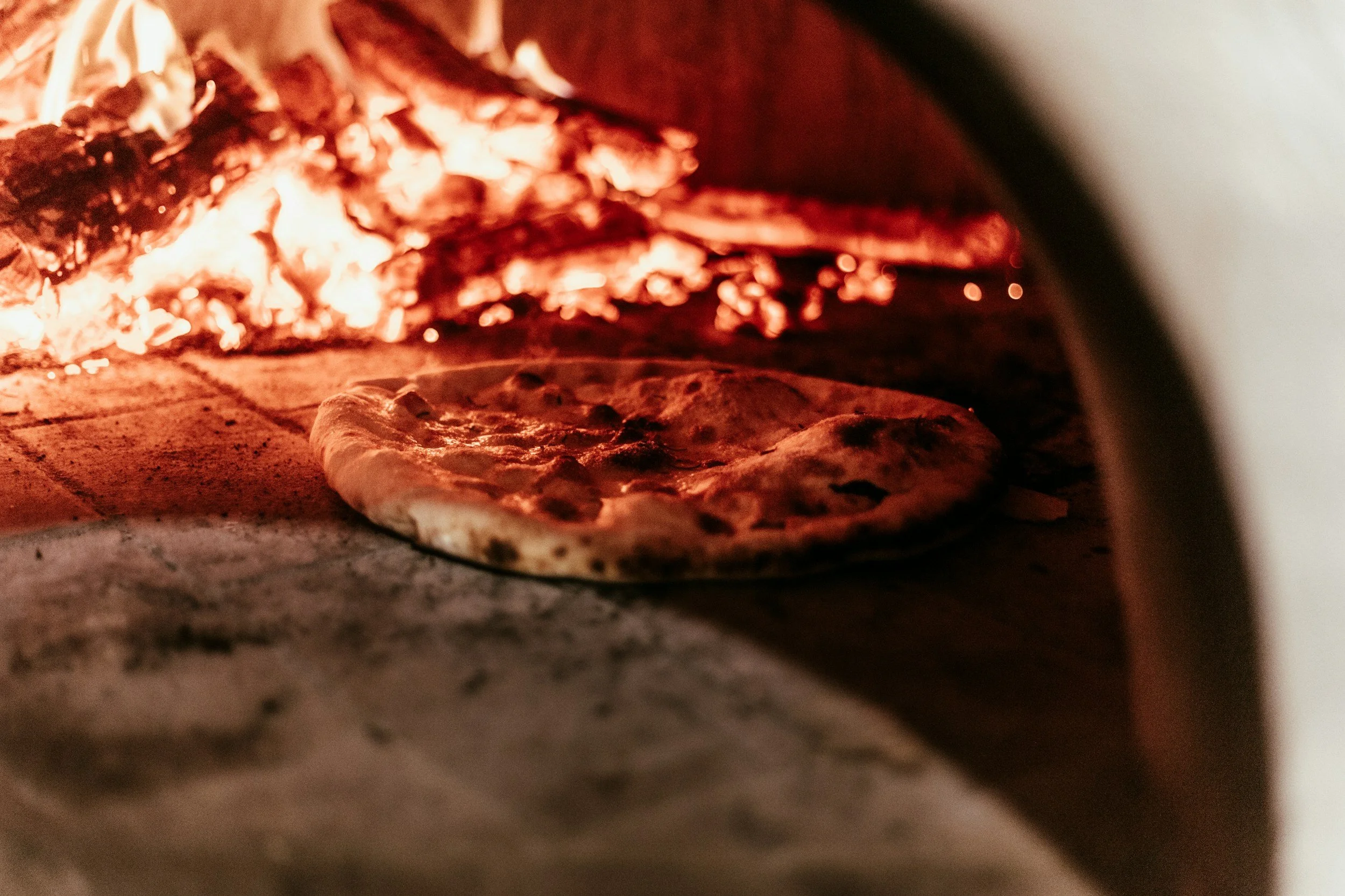 A pizza baking inside an oven with glowing red heat, visible flames, and a stone surface beneath.