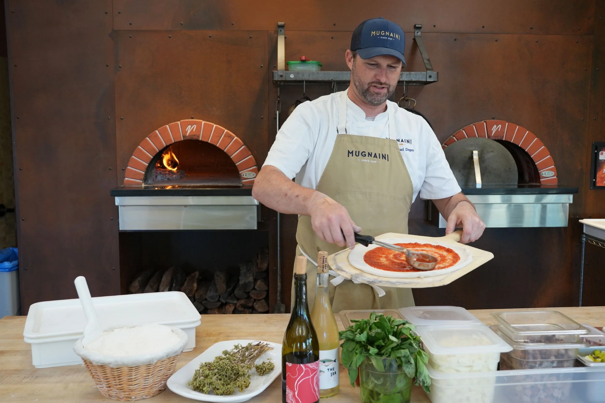 Man preparing a pizza in front of a wood-fired oven, with ingredients and bottles on the counter.