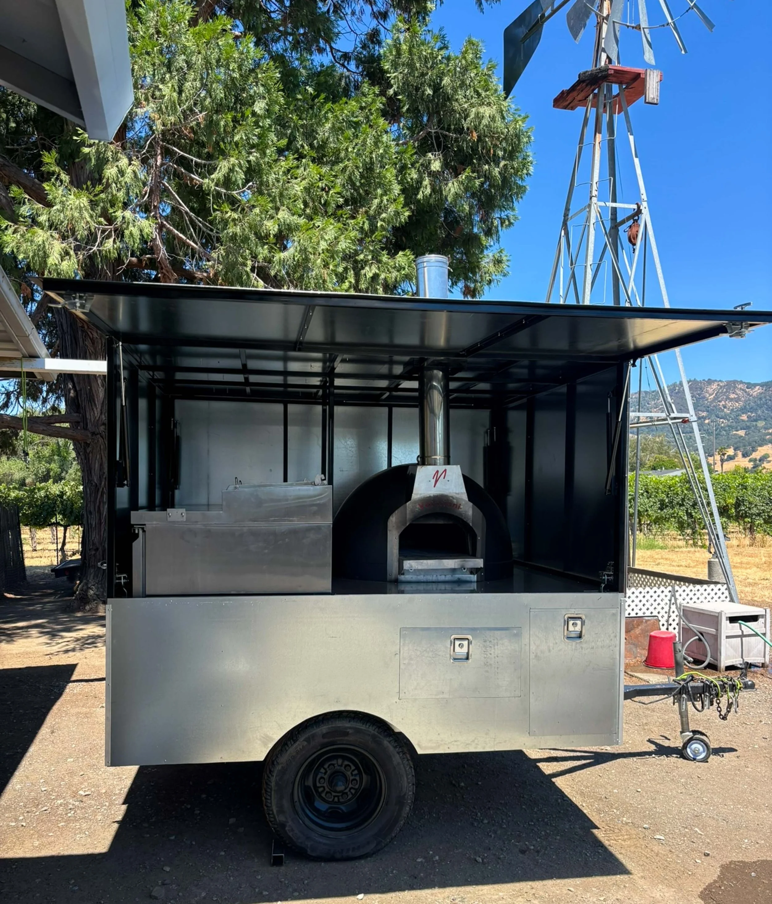 A mobile outdoor pizza oven trailer with a black pizza oven, stainless steel sink and counter, set up under a canopy with trees and a windmill in the background.