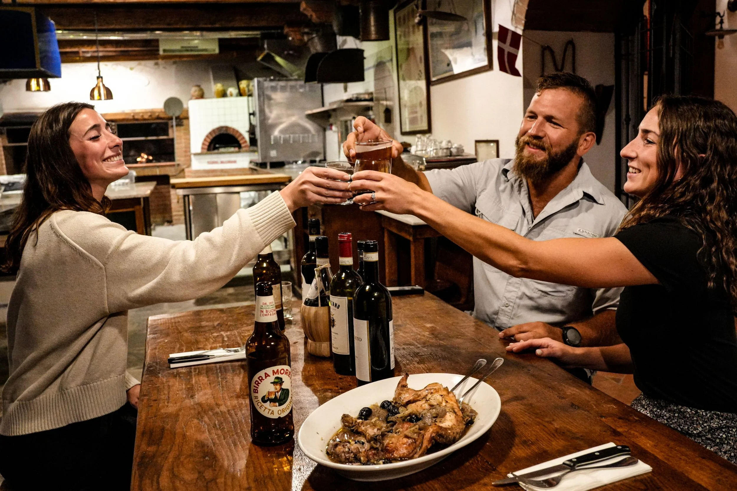 Three people at a restaurant celebrating with drinks, toasting over a wooden table with a roasted chicken dish, wine bottles, and beer bottles, smiling and enjoying each other's company.