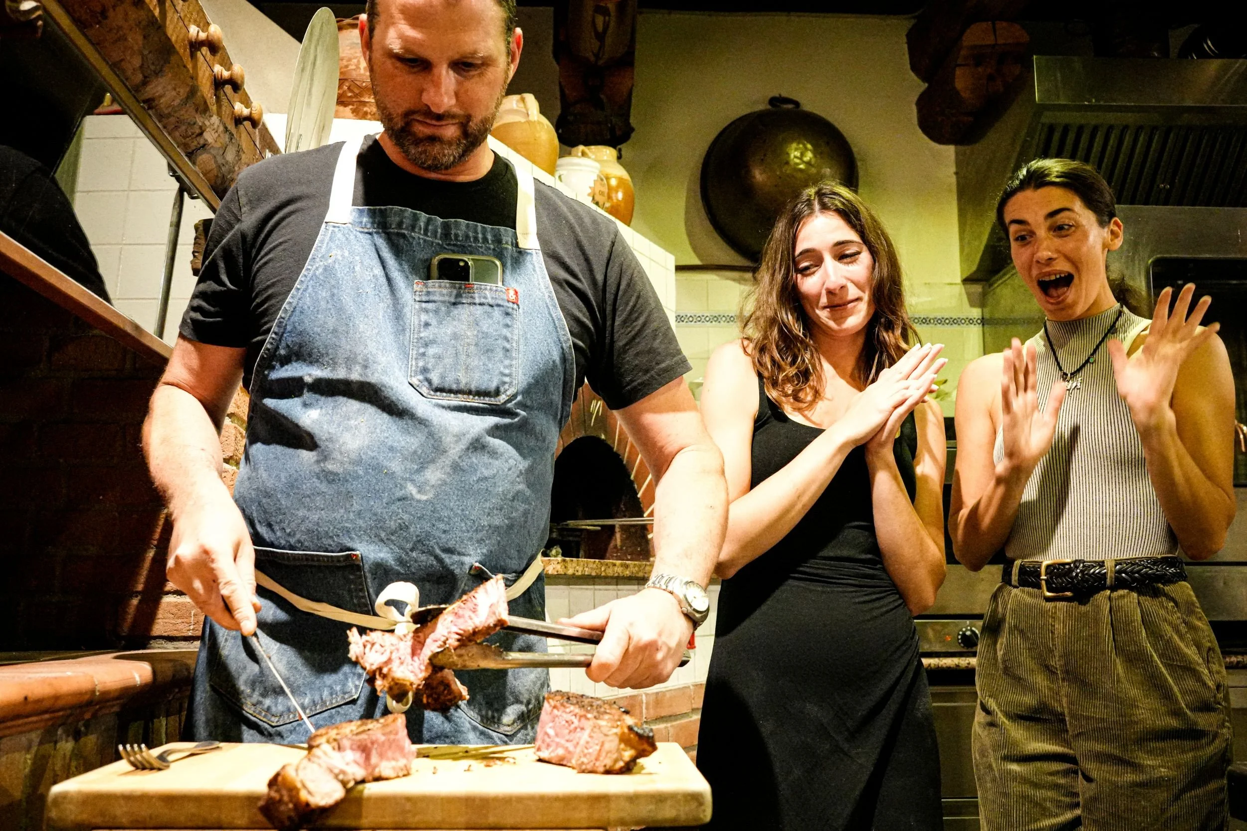 Chef Degen  cuts a piece of cooked steak on a wooden cutting board, while two guests are watching and clapping in a kitchen environment.