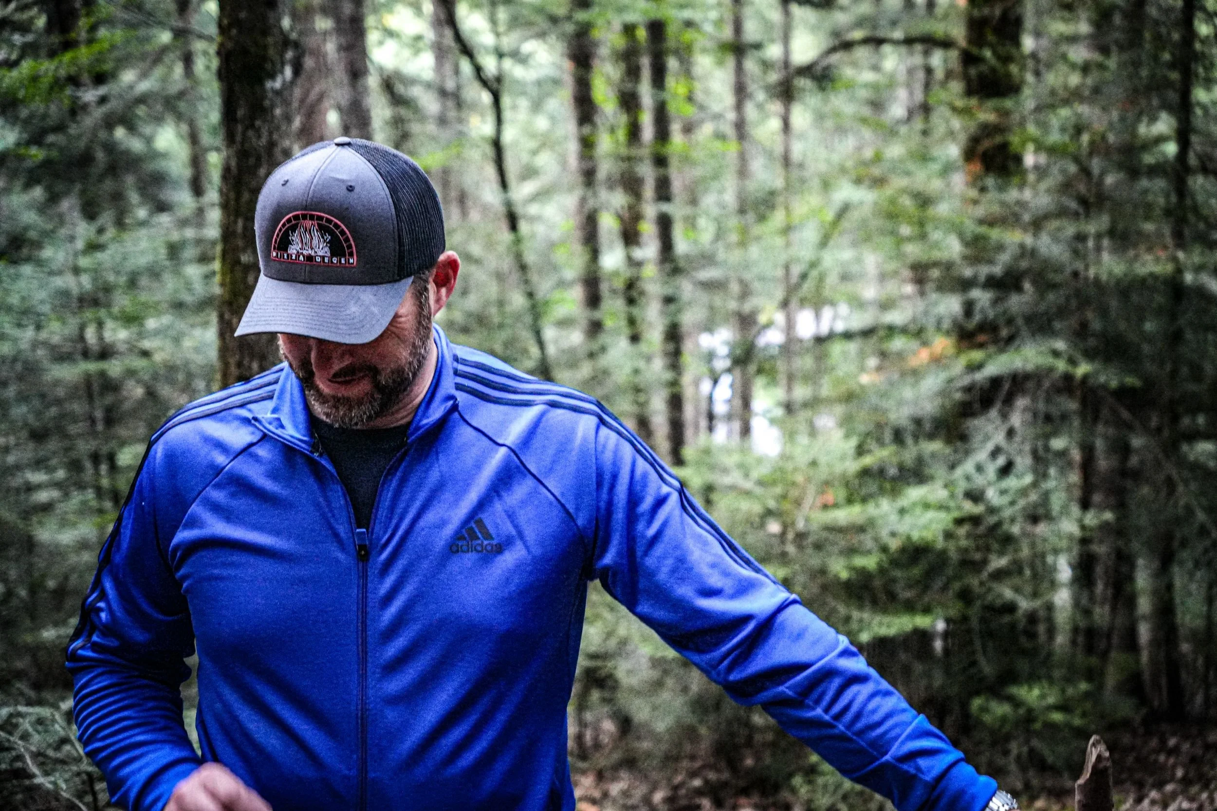 A man in a blue Adidas jacket and a black and gray cap walking through a forest with green trees.