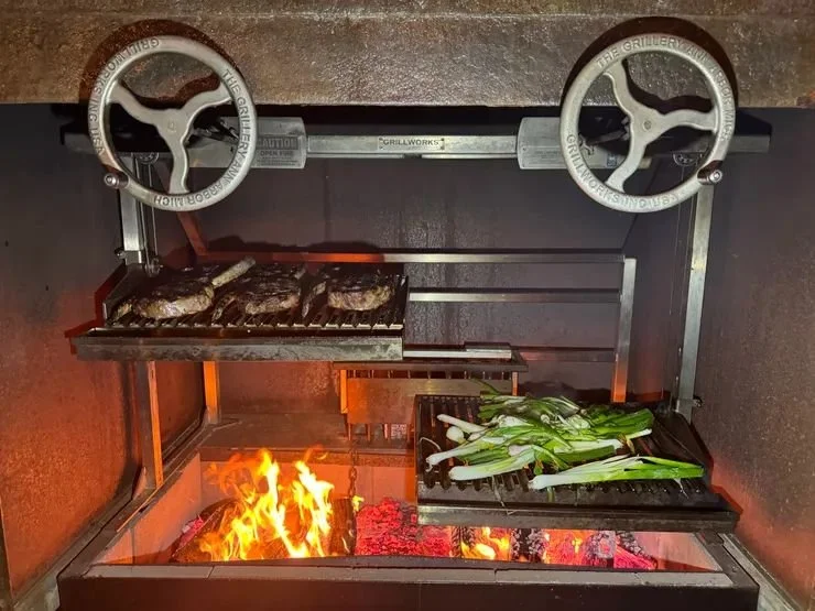 Inside a grill, meat patties are cooking on the upper rack, and green onions are grilling on the lower rack. There is a small fire with glowing coals below.