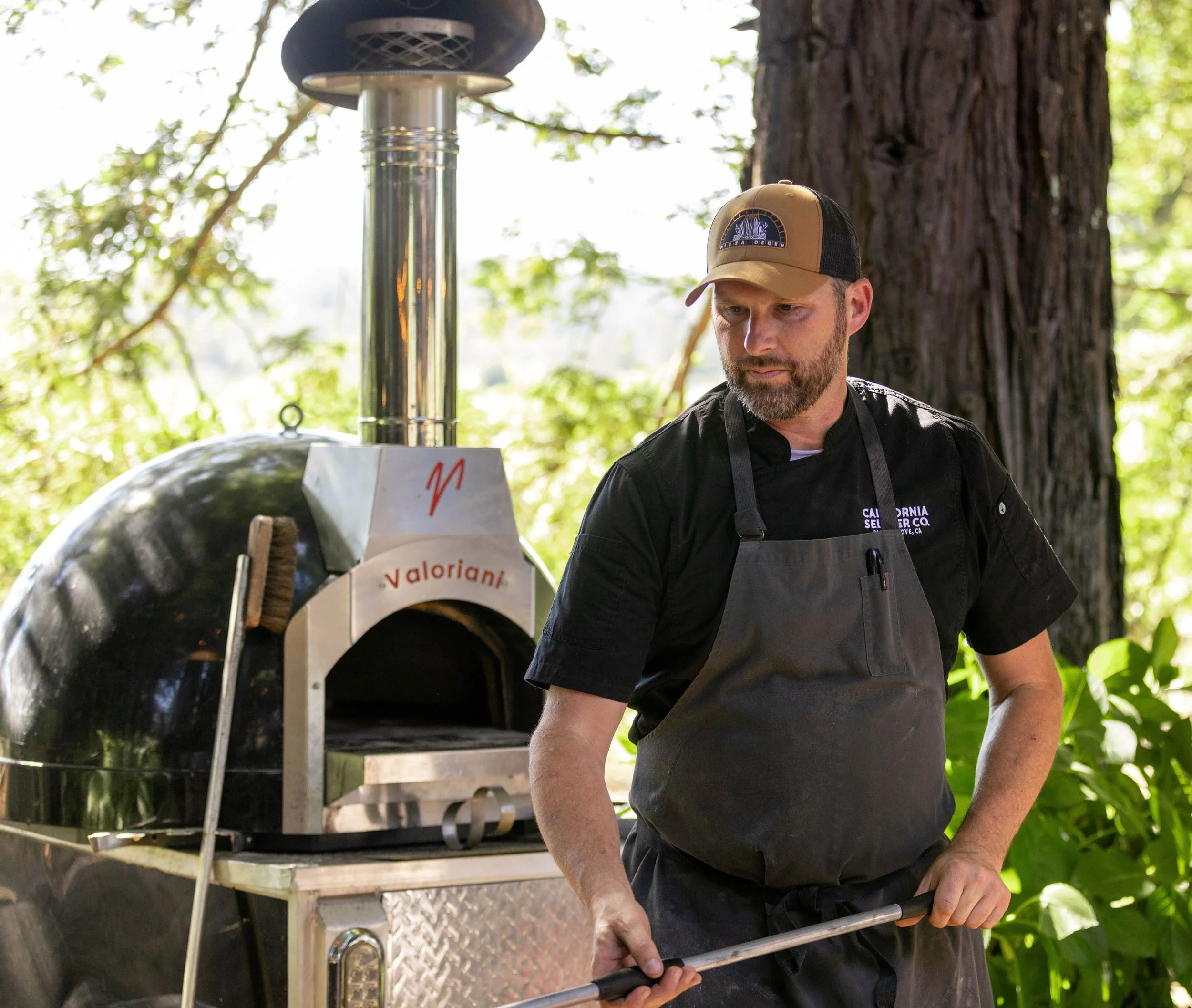 A man wearing a black chef's uniform and a brown cap stands outdoors next to a wood-fired pizza oven, surrounded by green trees and foliage.