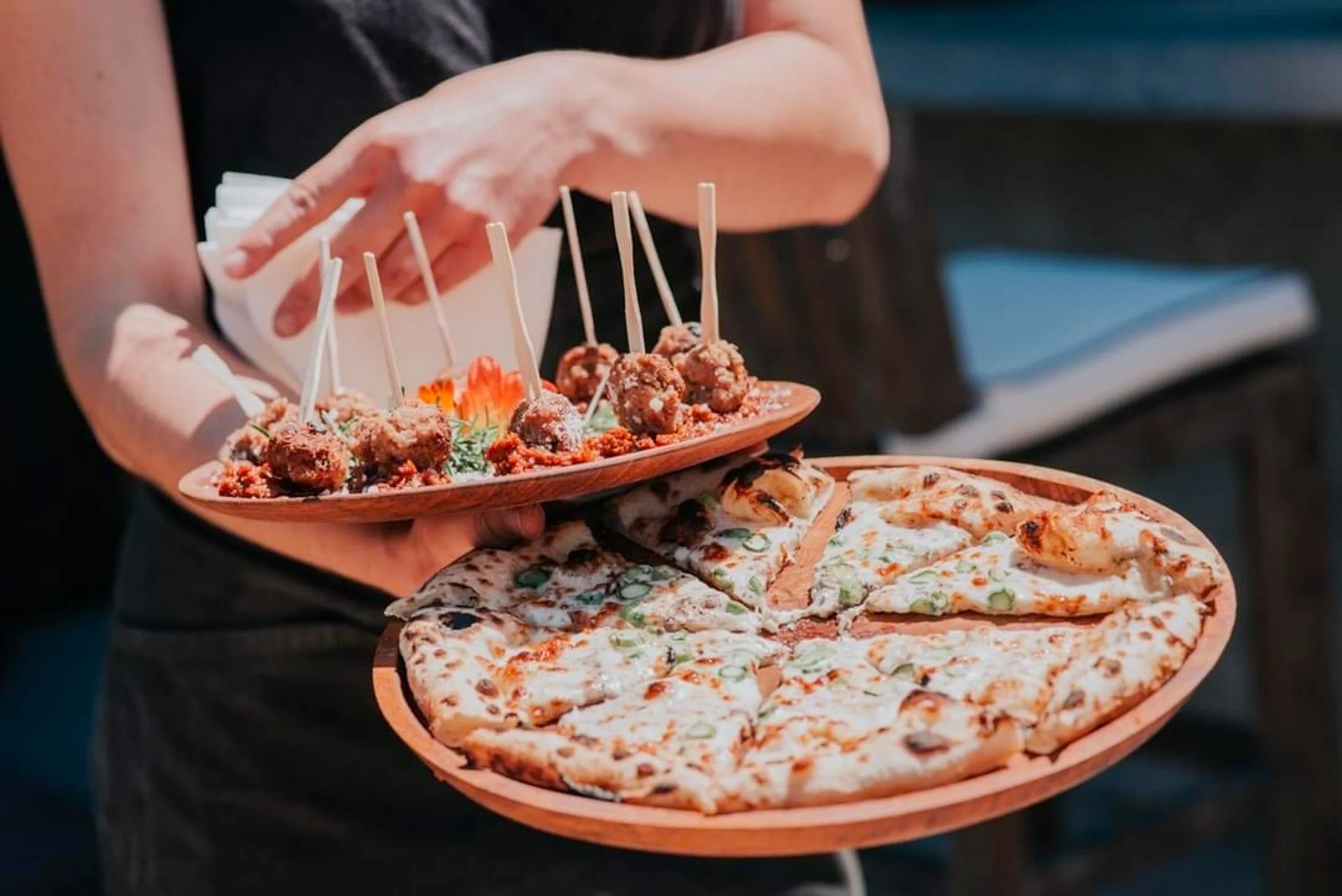 Person holding two wooden plates with food: one with a pizza and the other with fried appetizer skewers.