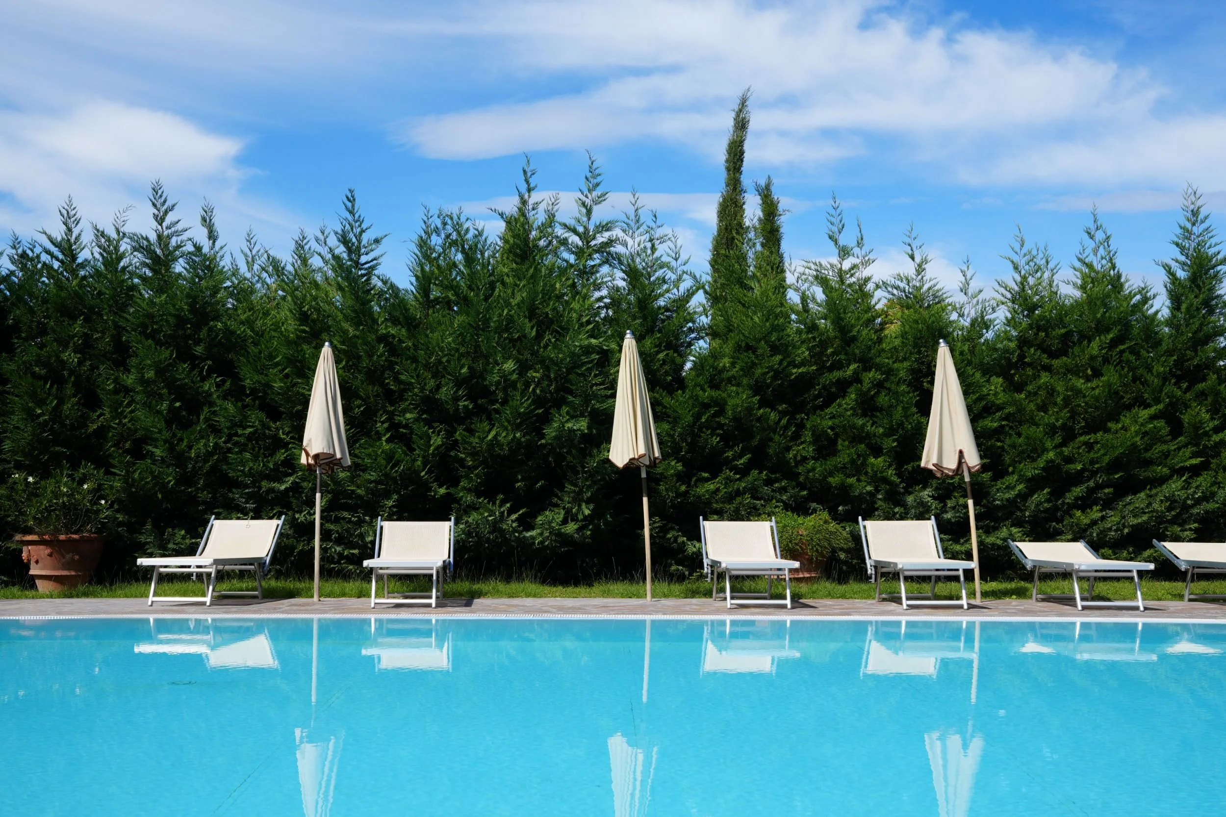 Empty poolside area with loungers and umbrellas, green hedge background, blue sky with clouds.