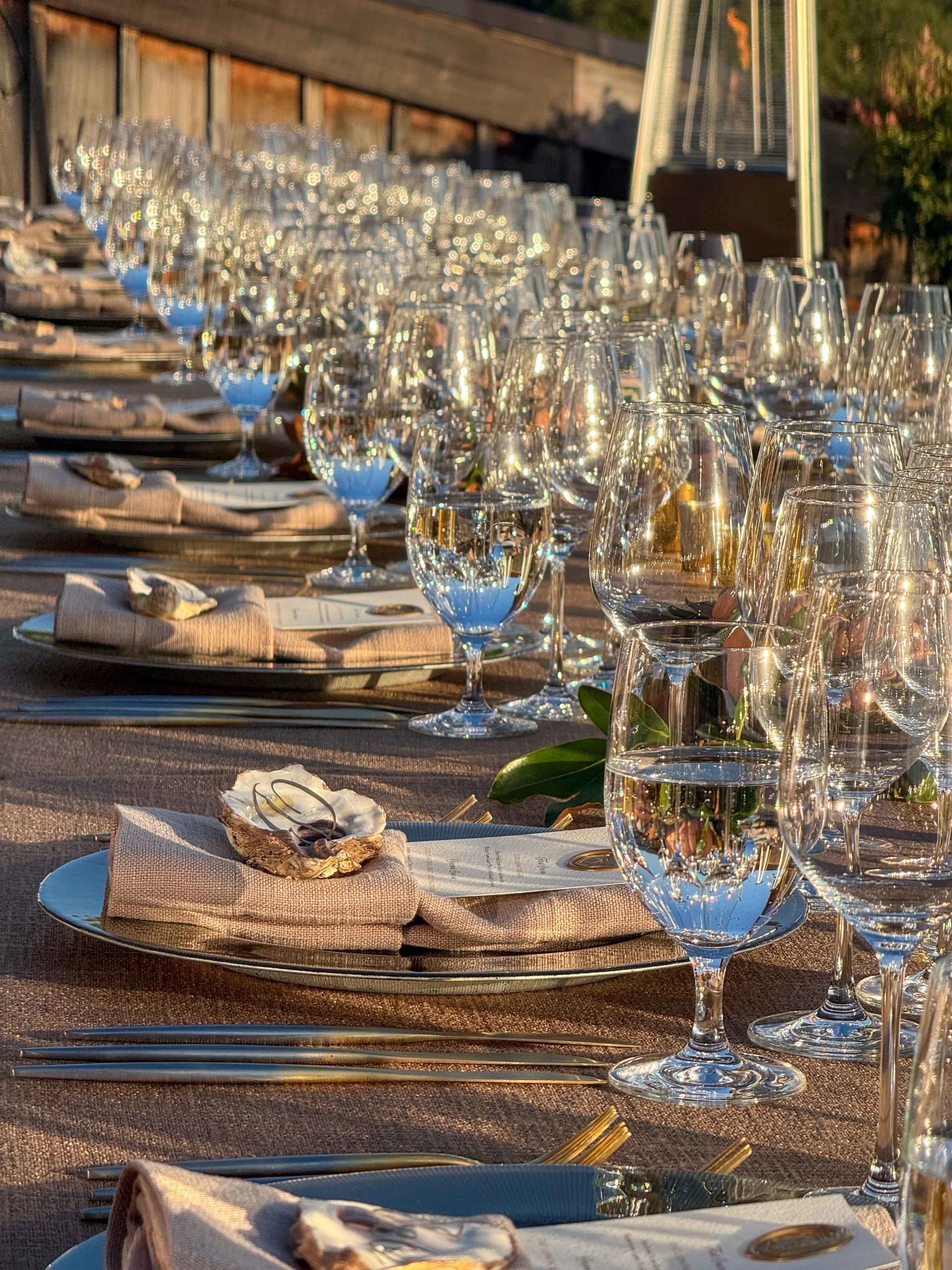 A long outdoor dining table set with multiple wine glasses, water glasses, plates, napkins, and shells with jewelry on top, illuminated by warm sunlight.