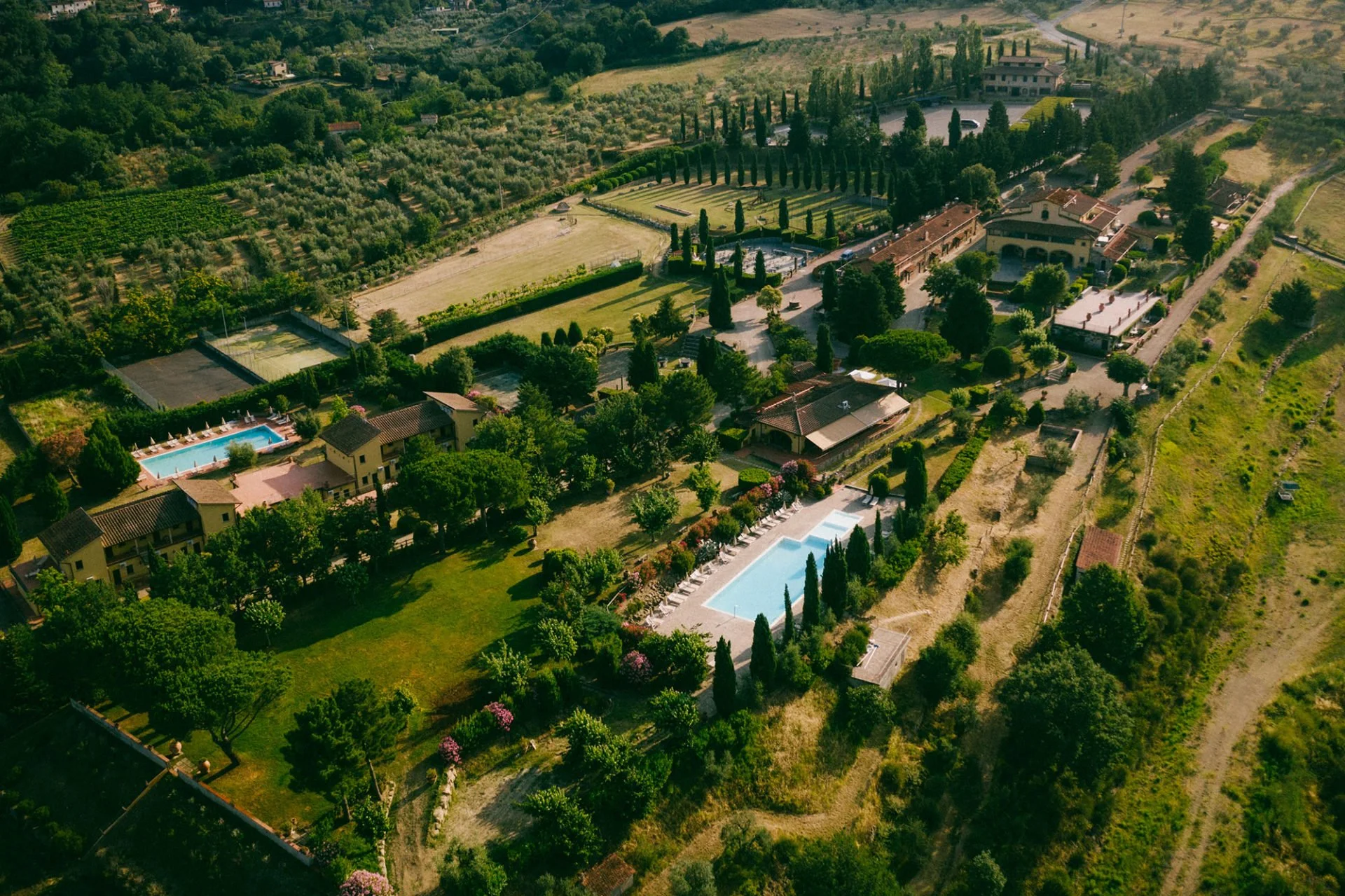 Aerial view of a large estate with multiple swimming pools, surrounded by lush greenery, trees, and landscaped gardens, with tennis courts and buildings in the background.