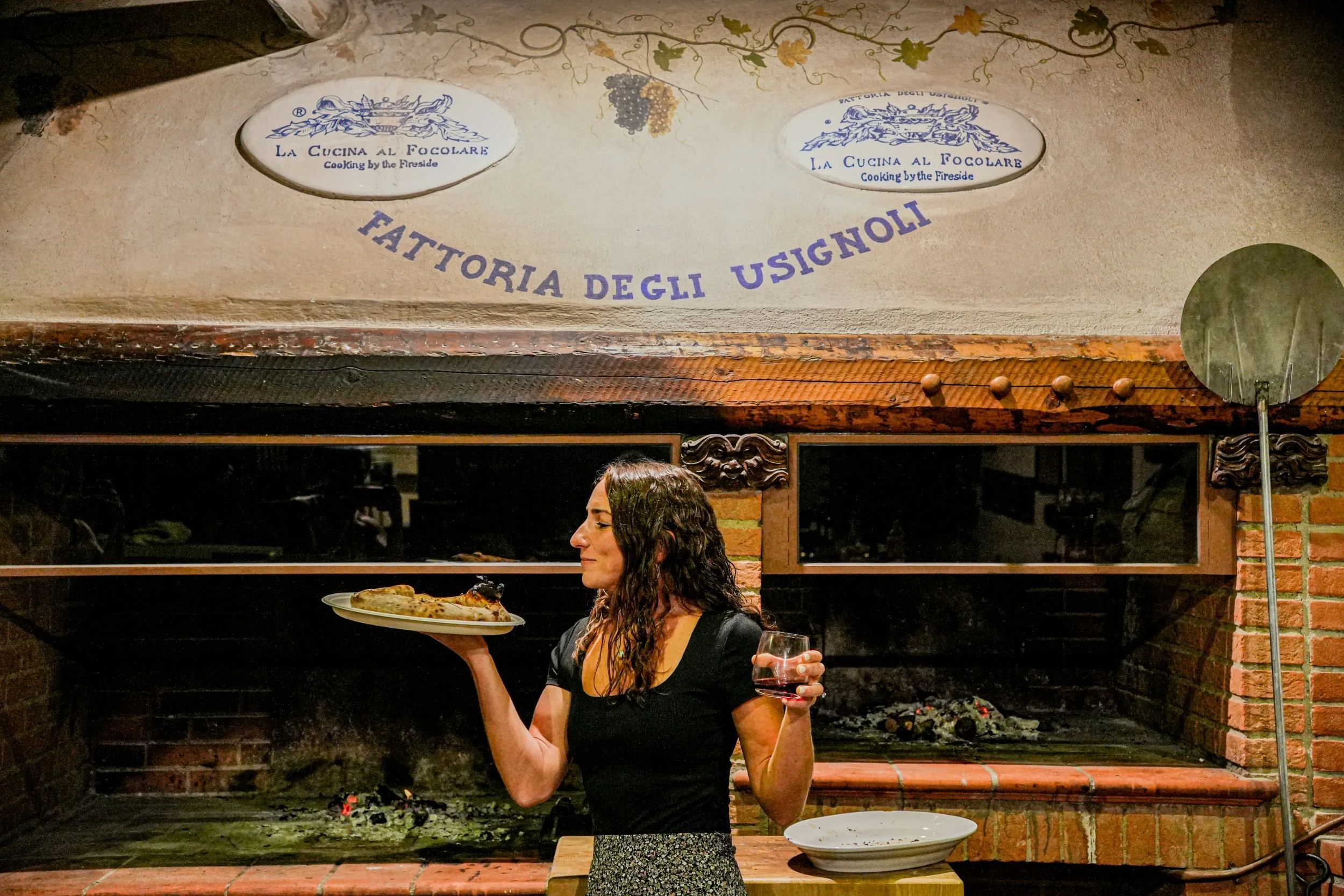 A woman standing in front of a brick fireplace, holding a plate of food in her right hand and a glass of wine in her left, smiling with her eyes closed.