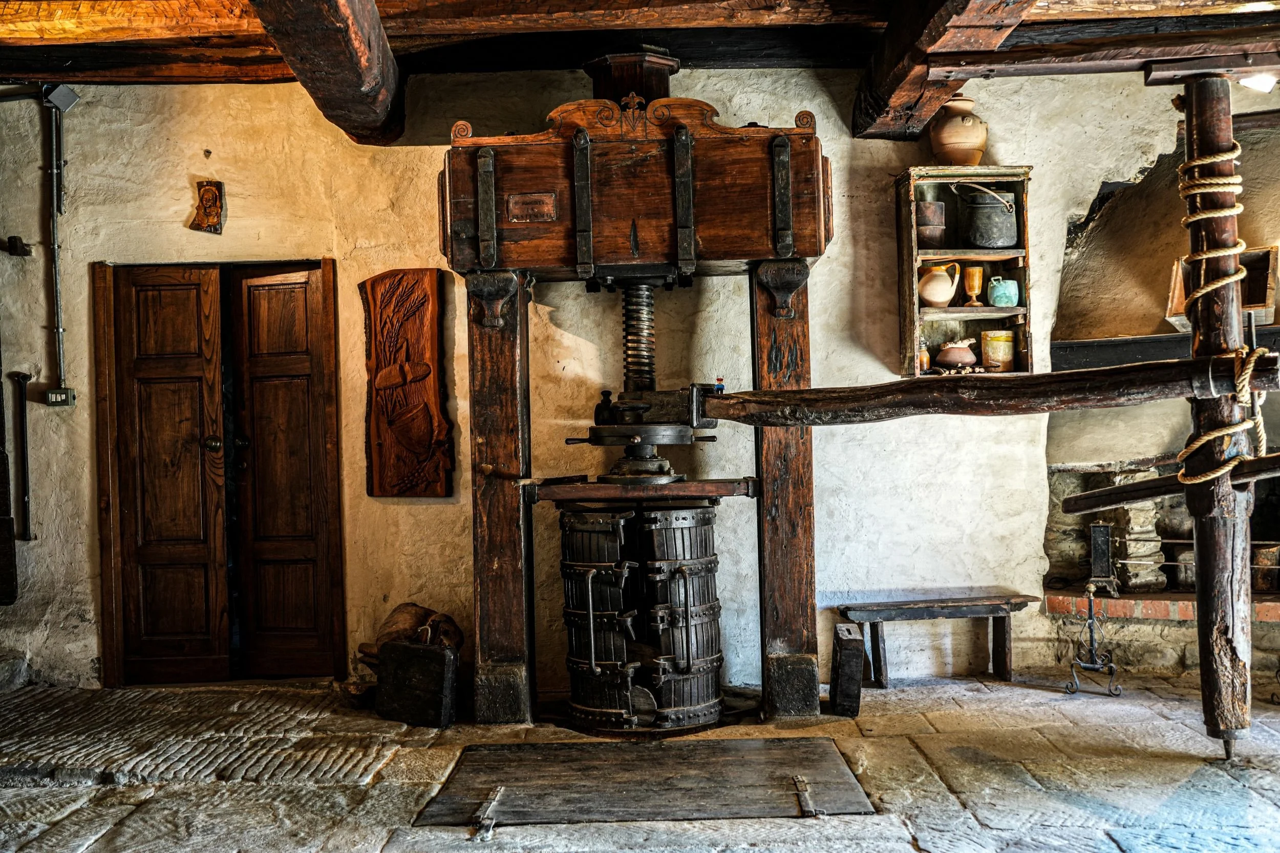 A historic interior with old wooden furniture and a large screw press at the center, surrounded by stone walls and rustic decor.