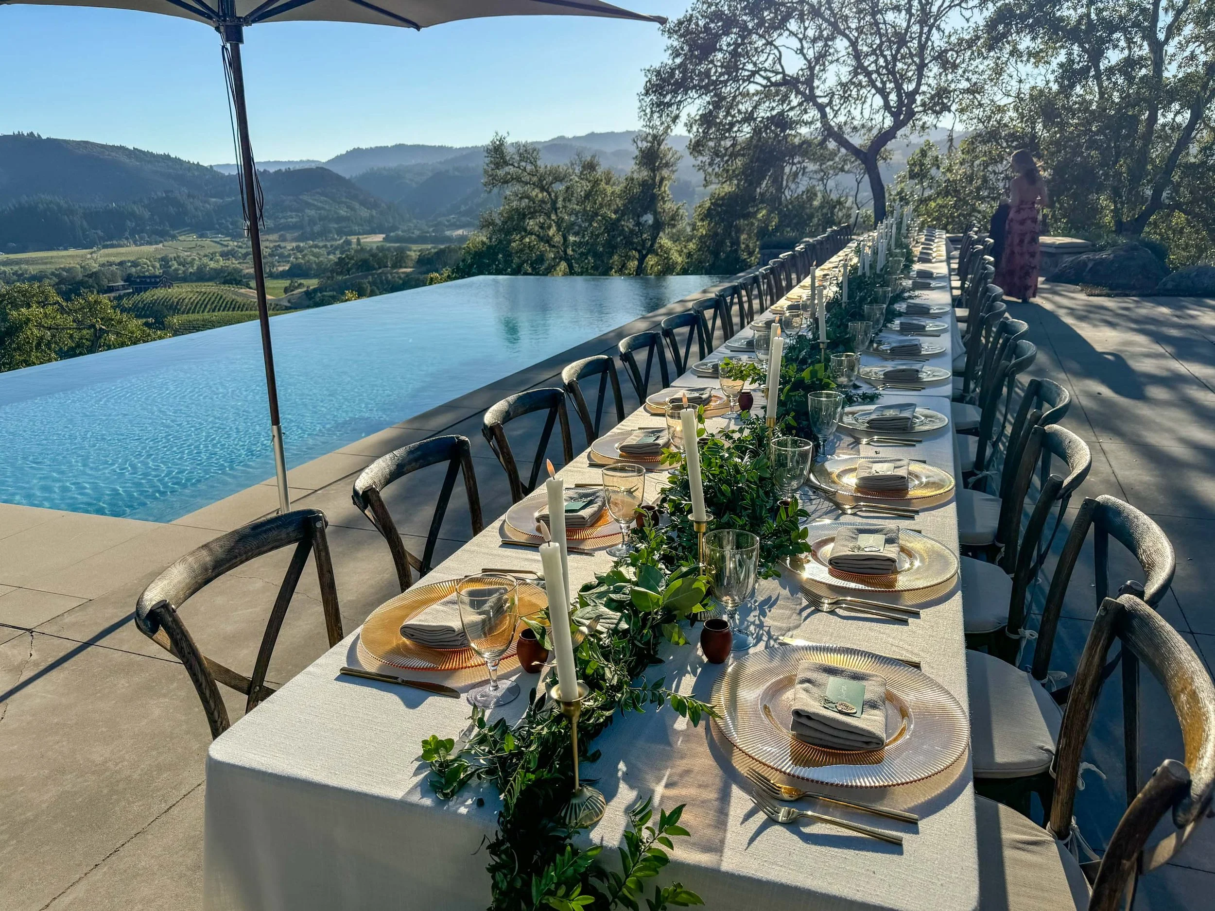 Long rectangular outdoor dining table set for a formal meal with plates, glasses, napkins, and candles, surrounded by chairs, overlooking an infinity pool with a scenic valley and hills in the background.