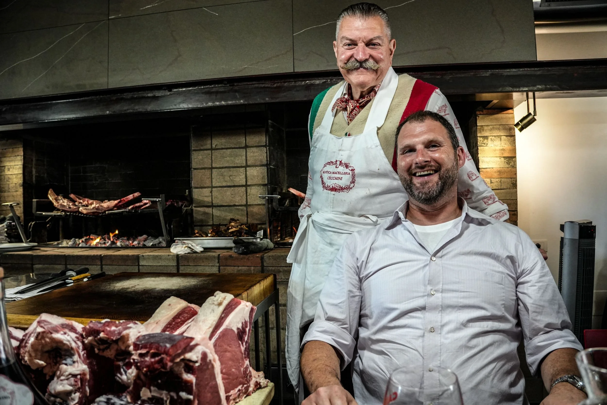 Two men smiling, one seated and one standing in a restaurant, with a brick fireplace and grill in the background, and raw meat on a cutting board in the foreground.