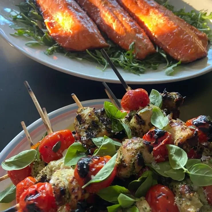 Close-up of a plate of grilled cherry tomato and basil skewers with melted cheese, in the foreground, and a plate of glazed salmon fillets garnished with greens in the background.