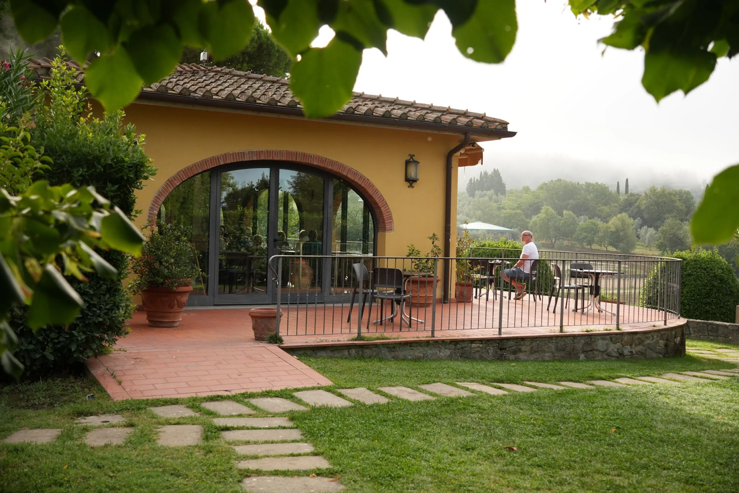 A person sitting alone on a terrace outside a yellow house with a large arched window, surrounded by greenery and trees, with a misty landscape in the background.