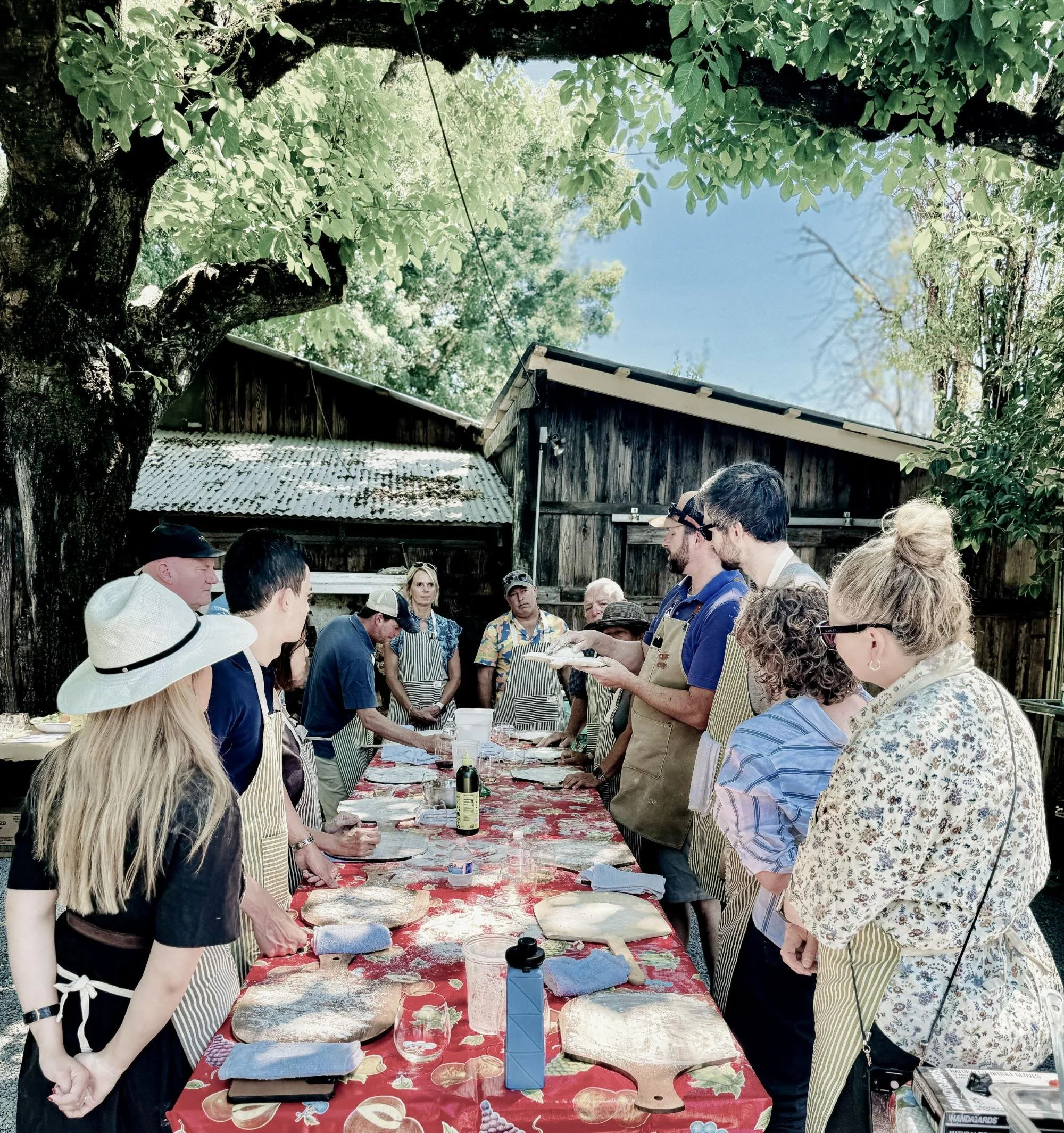 People gathered around a long table outdoors, participating in a cooking class or demonstration, with an instructor in the center explaining something, surrounded by trees and rustic wooden buildings.