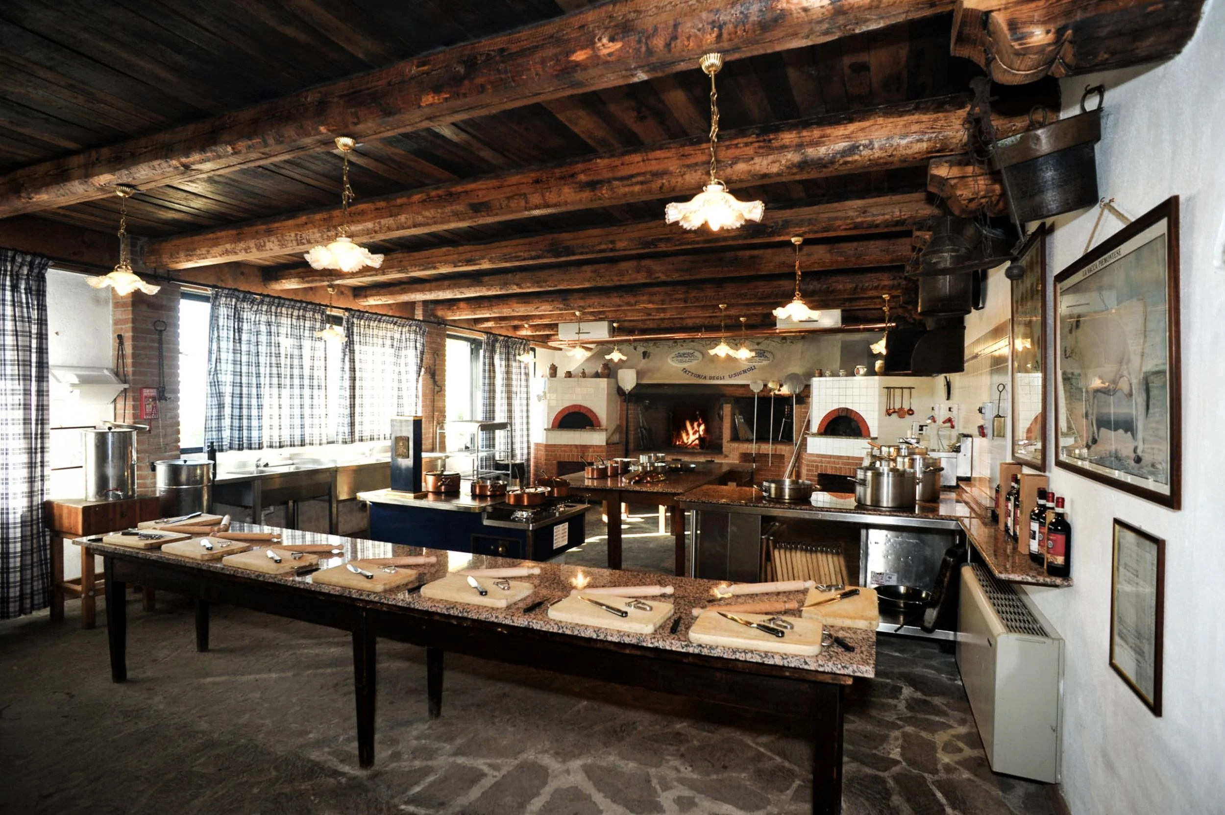 Interior of Italian restaurant with wooden ceiling beams, checkered curtains, and a stone floor. Features a cooking area with copper pots, a fireplace with a fire, and framed pictures on the walls.