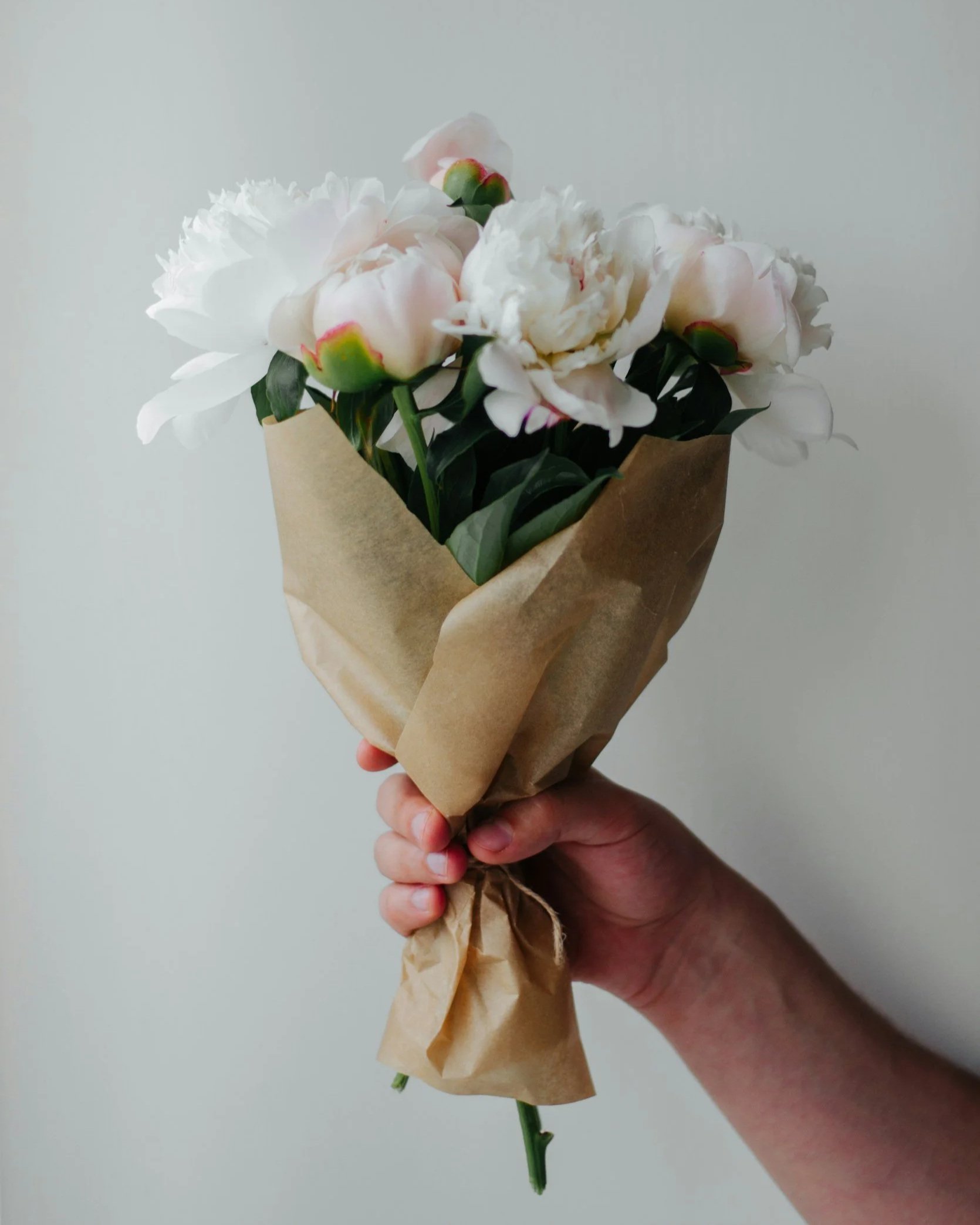 Hand holding a small bouquet of white peonies wrapped in brown paper