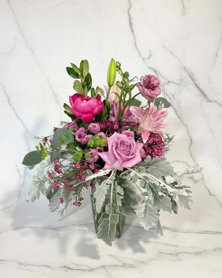 Pink and purple flowers in a clear glass vase against a white marble background, including roses, lilies, and other filler flowers by Dallas wedding florist Izalena Floral House.”