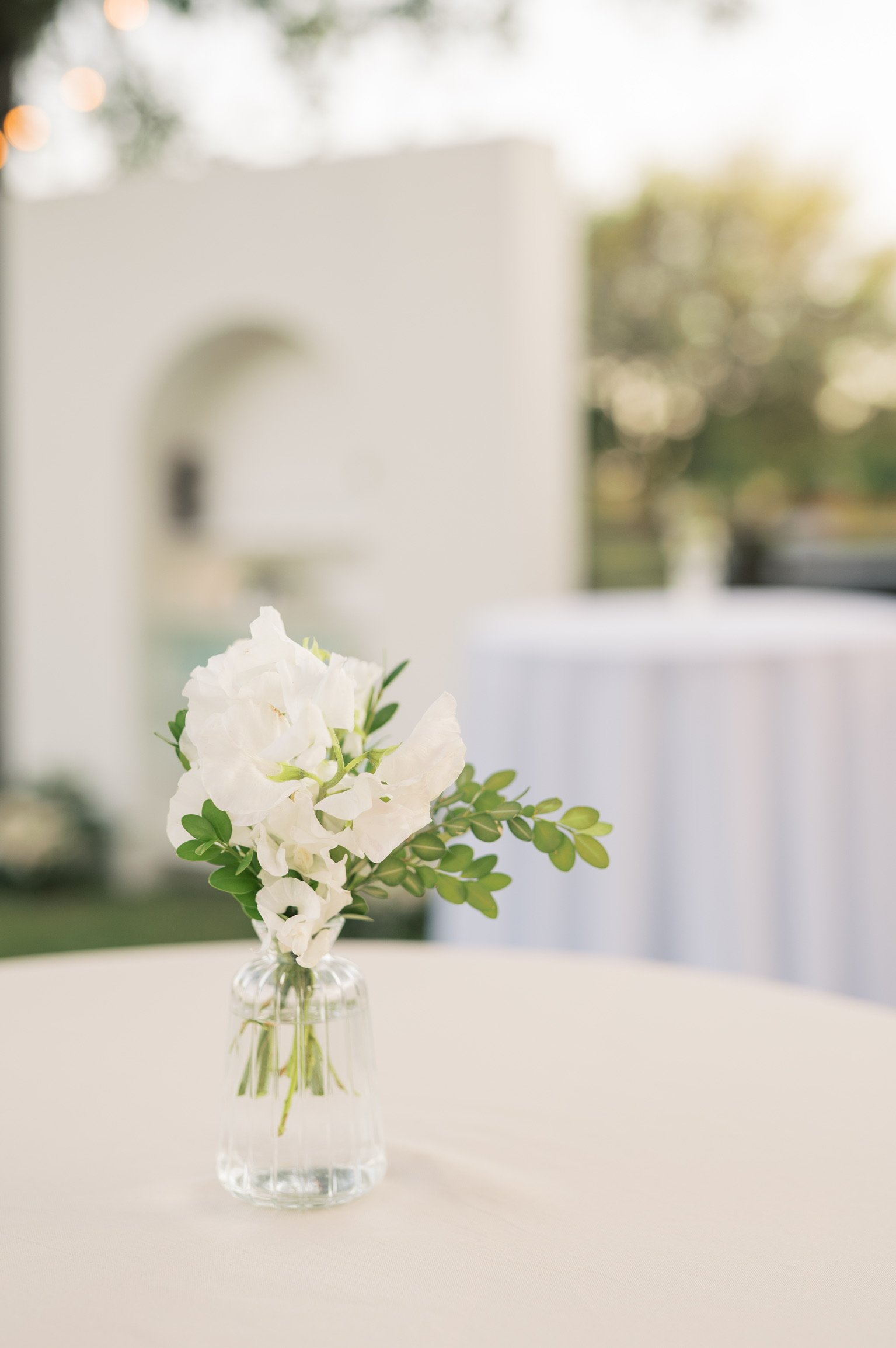 A small clear glass vase with white flowers and green leaves on a white table, with a blurry background of trees and natural light.