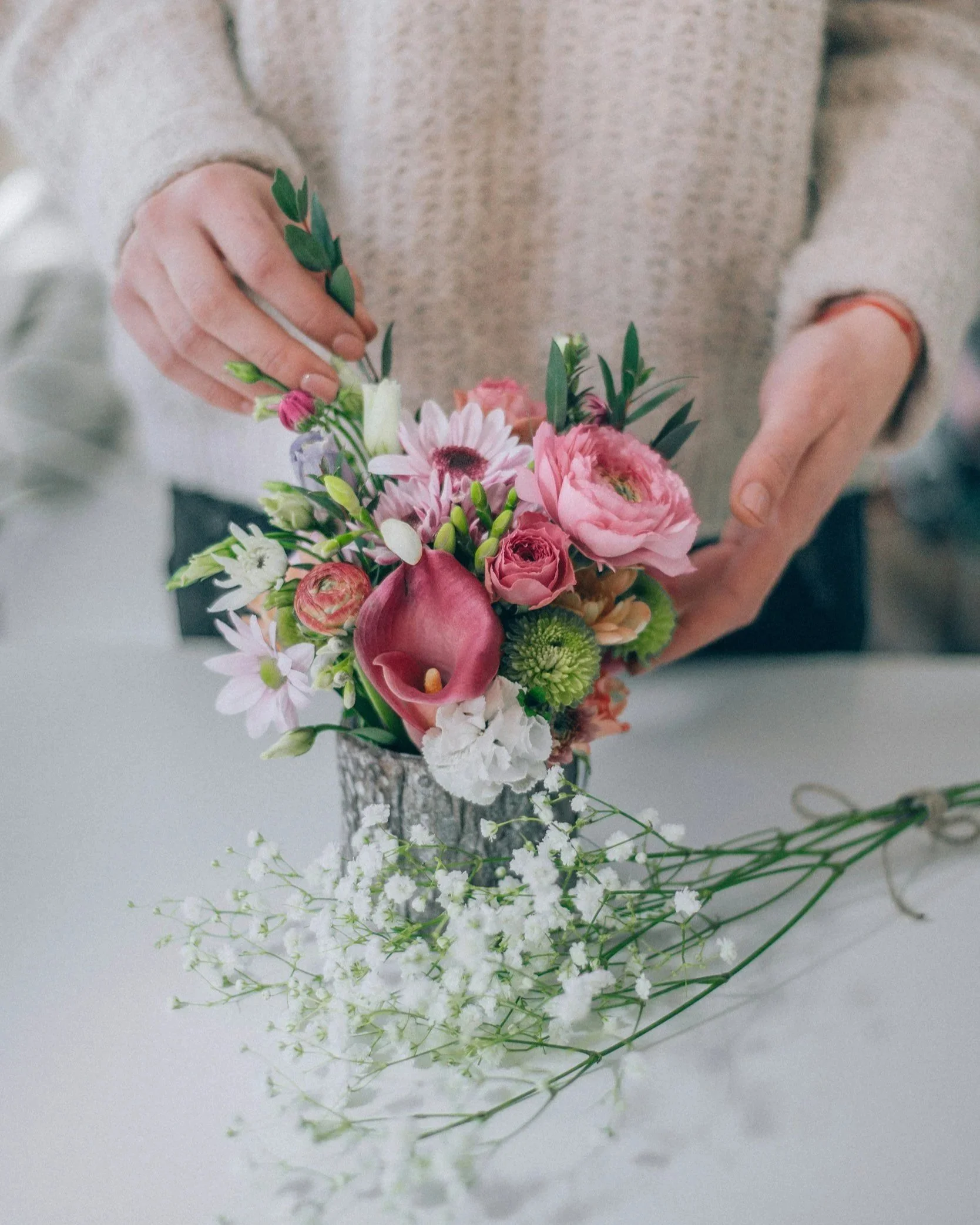 A hand arranging a bouquet of pink, white, and green flowers in a glass vase, with a soft sweater worn by the person by Dallas wedding florist Izalena Floral House.