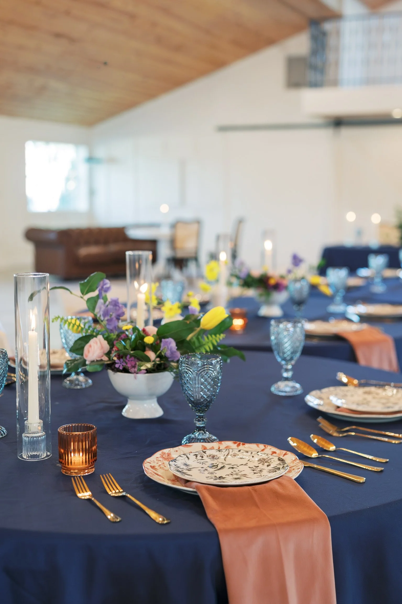 A dining table decorated for a celebration with a centerpiece of pink, purple, yellow, and green flowers in a white bowl, blue tablecloth, gold utensils, patterned plates, candles in glass holders, and a pink napkin. In the background, part of a room