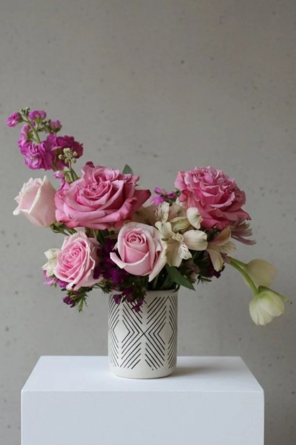A bouquet of pink roses, white flowers, and purple flowers in a white geometric-patterned vase on a white pedestal against a plain gray wall by Dallas wedding florist Izalena Floral House.