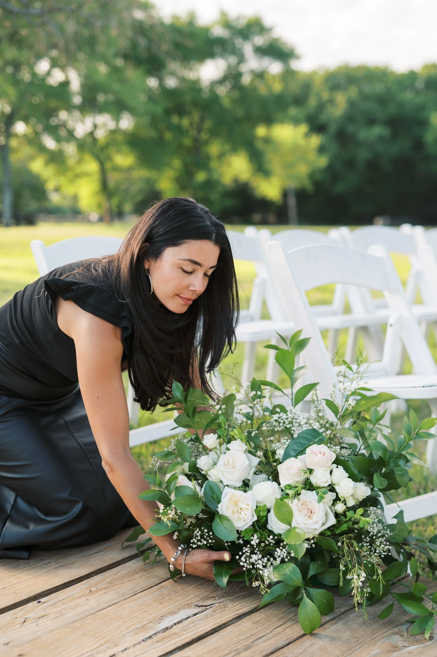 A woman arranging a floral centerpiece with white roses and greenery outdoors with white chairs and trees in the background.