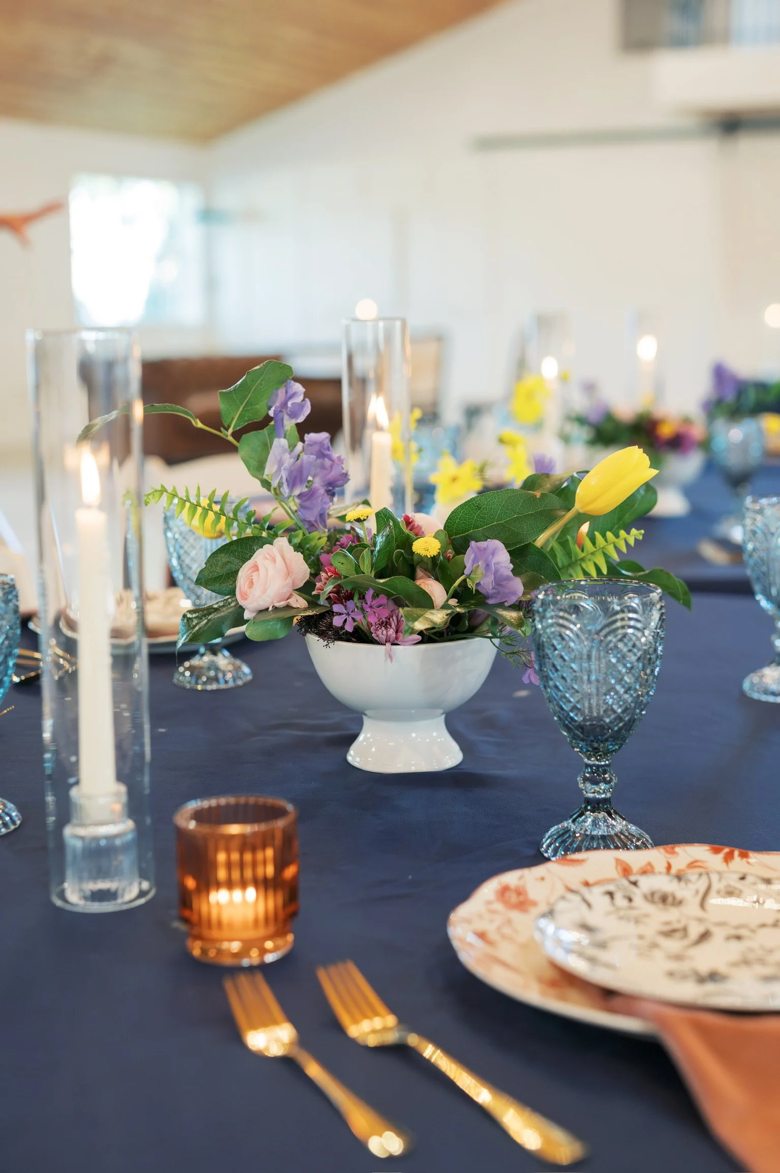 A table decorated for a special occasion with a dark tablecloth, a floral centerpiece with purple, yellow, and pink flowers, and lit candles. Place settings include patterned plates, gold utensils, and glassware, with some glasses in amber and textured blue designs.